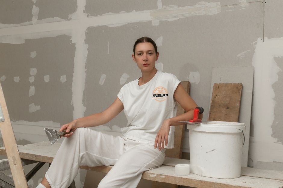 A woman in white work clothes sits on a bench in a room under construction, holding a drywall tool. Surrounded by tools and drywall, she is a drywall contractor near N 58th Pl, and E Vía Del Cielo, Paradise Valley, AZ.