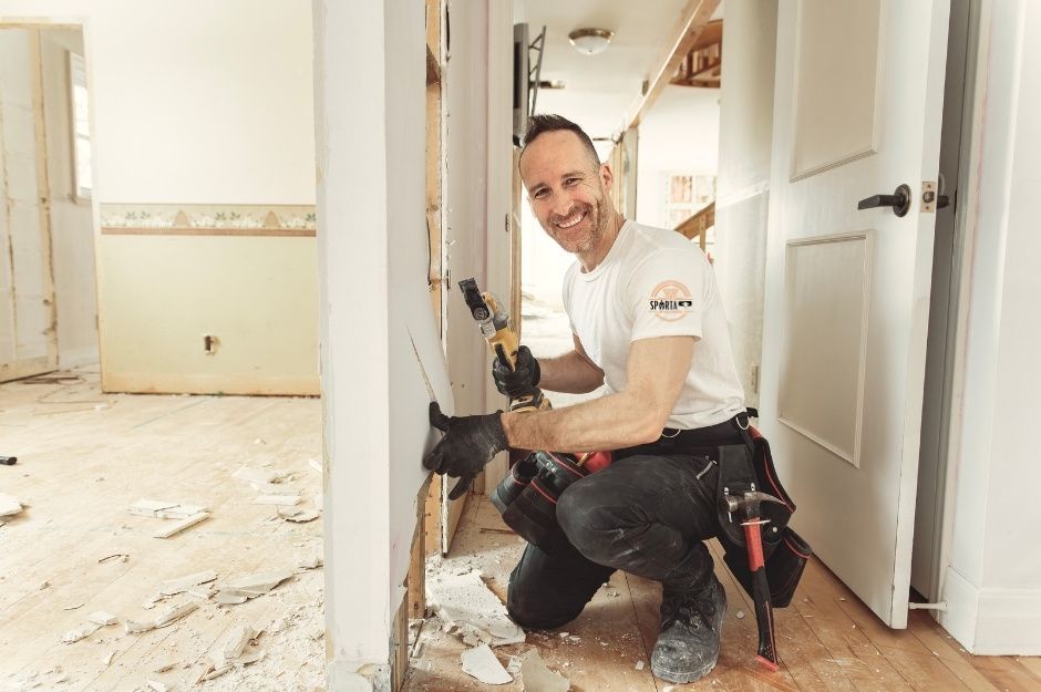 A drywall contractor in a white t-shirt kneels while using a power tool for drywall installation in a room with white walls and scattered debris on the floor. This drywall company professional is working on a project near E Bonanza Way, and S 215th St, Queen Creek, AZ.