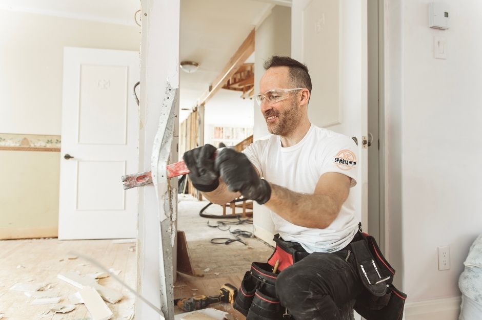 A professional drywall contractor in a white t-shirt uses a power tool for drywall smoothing while kneeling among construction materials and white walls. This drywall company specialist is completing work near E Rock Rd, and N Dolomite Ln, San Tan Valley, AZ.