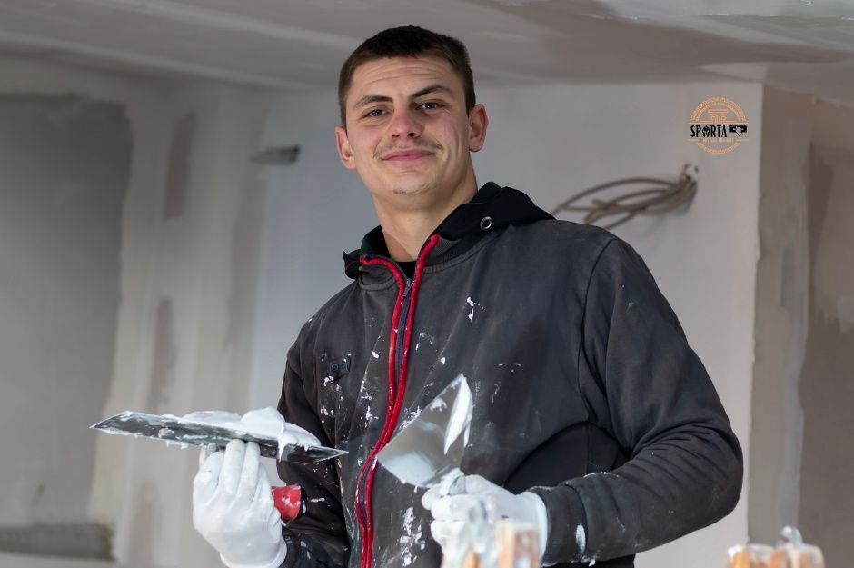 A drywall contractor wearing a dark gray hoodie holds a drywall knife and joint compound while standing in a room with white walls and a ceiling. He is working on a project near E Bluejay Dr, and S Mesquite Trail, Spring Valley, AZ.