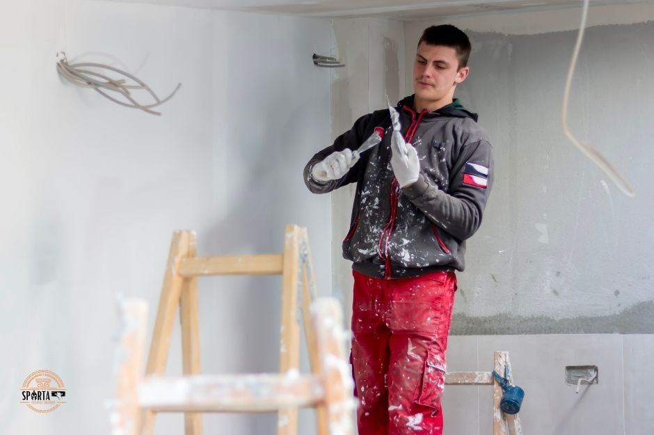 A drywall company residential construction worker in gray hoodie and red pants holds a putty knife in an unfinished room with white drywall walls and exposed ceiling fixtures. The professional is working on smoothing joint compound on wall surfaces near W Santa Ynez Dr, and W Los Bancos Dr, Sun City West, AZ.