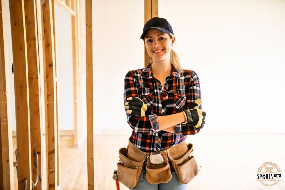 A drywall company worker stands confidently with arms crossed, wearing a plaid flannel shirt and hard hat with a tool belt around her waist. She is positioned in front of wooden wall framing in a residential construction site near E Copper Valley Ln, and E Minnesota Ave, Sun Lakes, AZ.