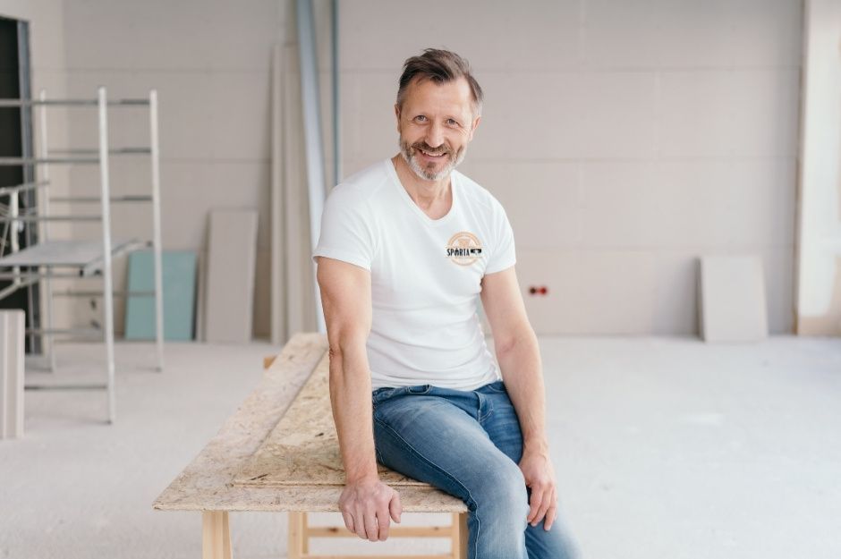 A drywall contractor in a white t-shirt and jeans sits on a wooden sawhorse in a room with freshly installed drywall walls and a ladder in the background. This professional contractor is working near E Velvet Dr, and S Taylor Dr, Tempe, AZ.
