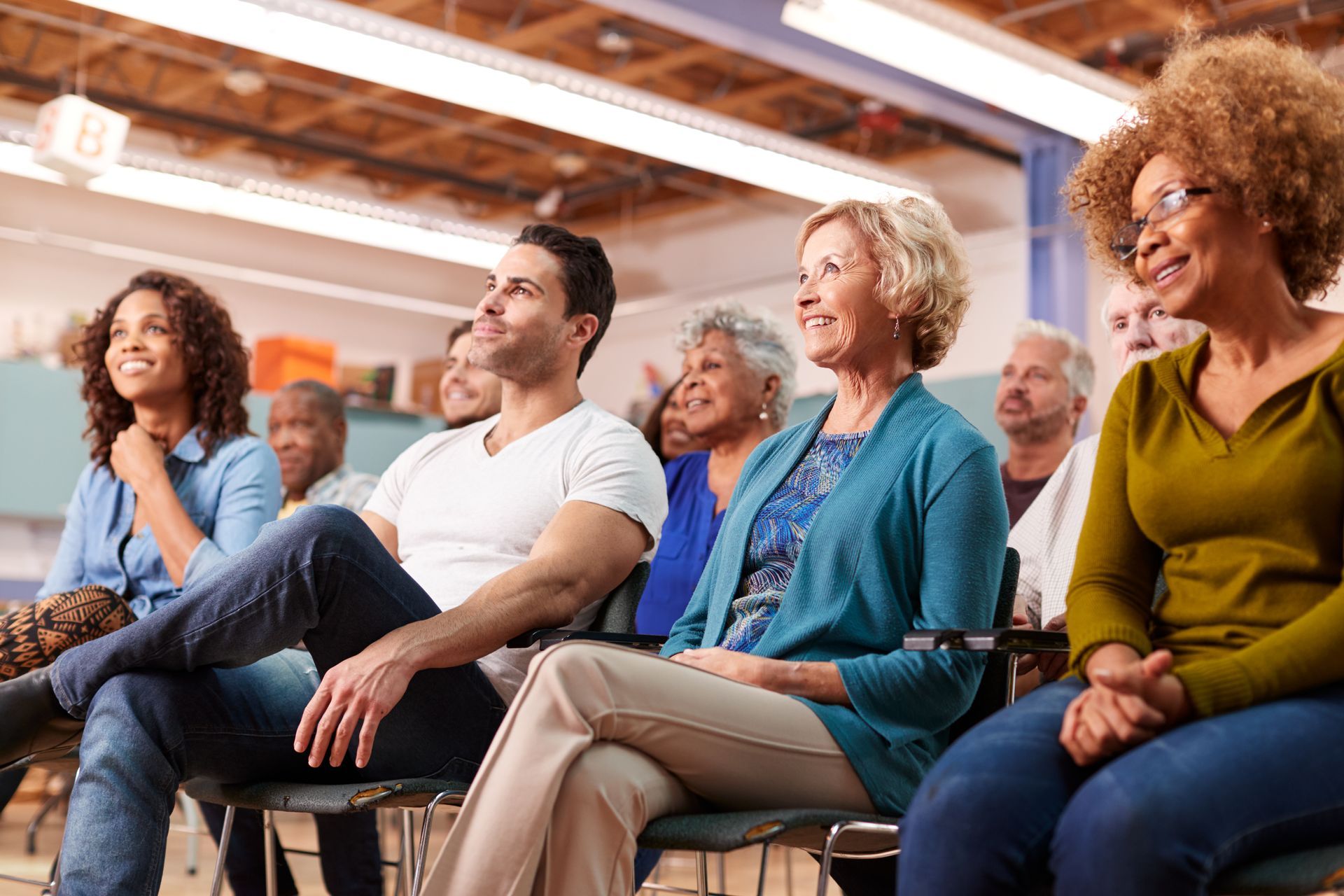 People seated, listening attentively in a brightly lit room. Diverse group with smiling faces and a focused atmosphere.