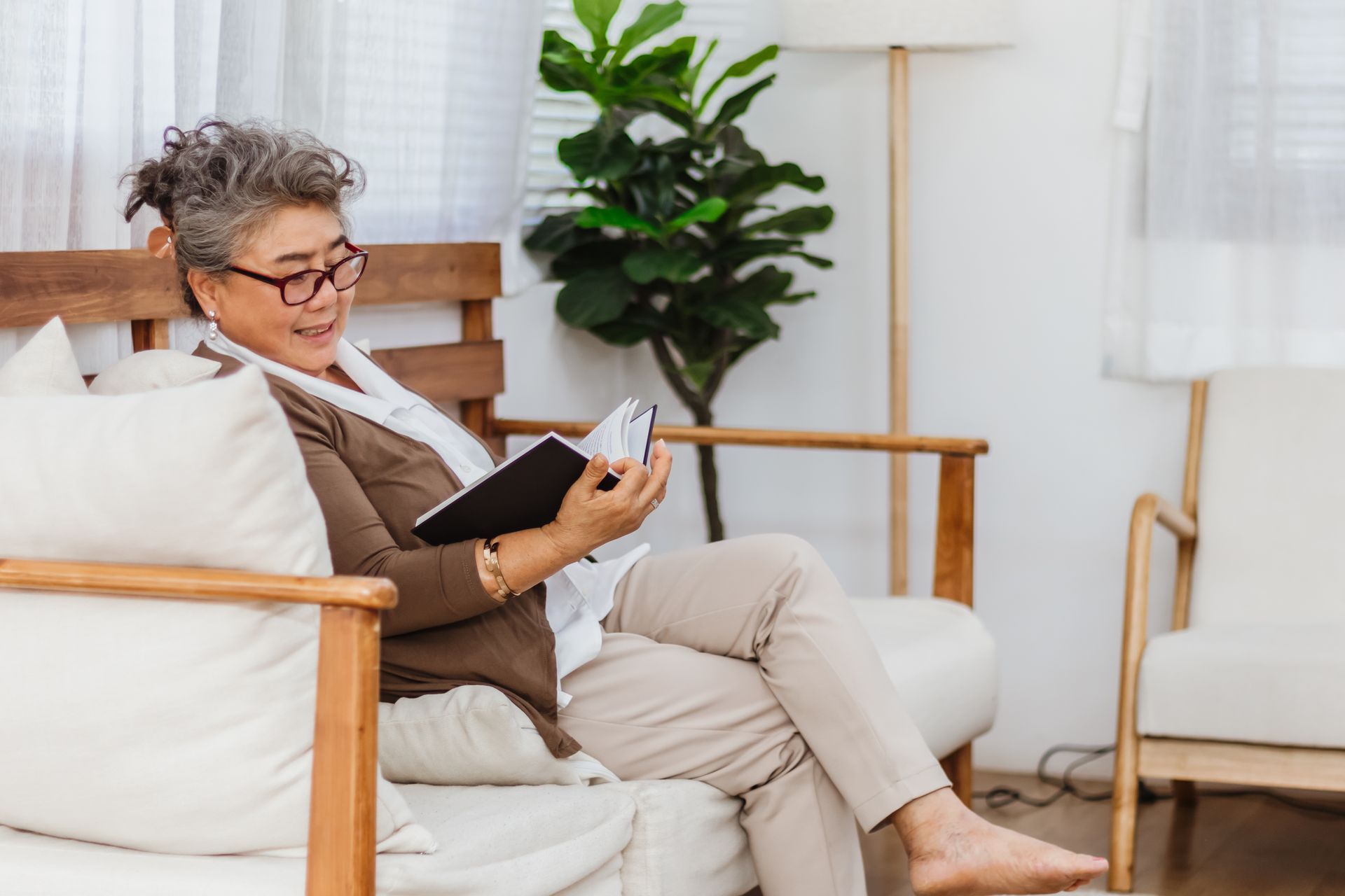 Senior Asian woman reading a book, smiling, in a bright living room.