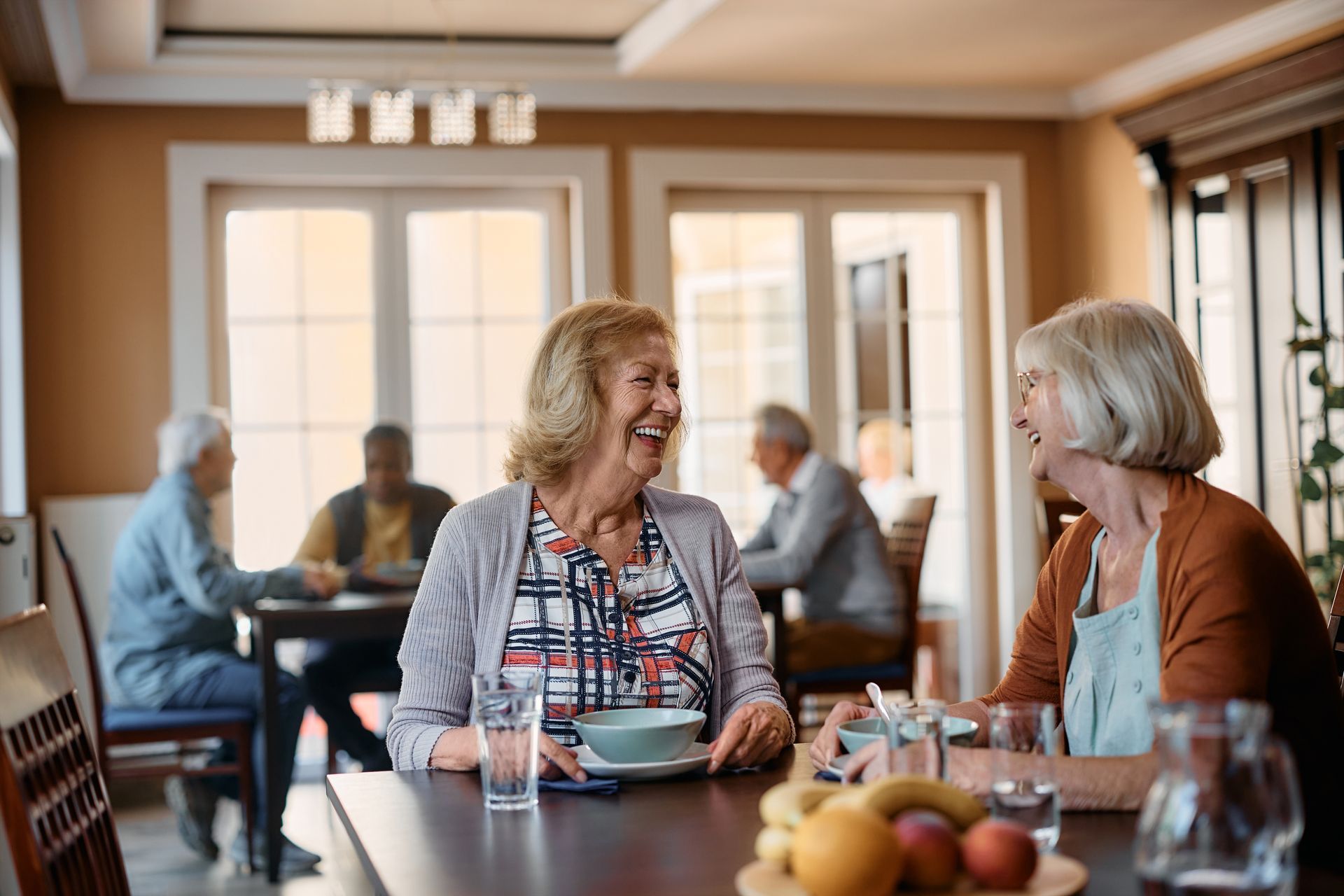 Two older women laugh at a table, dining in a brightly lit room with other residents.