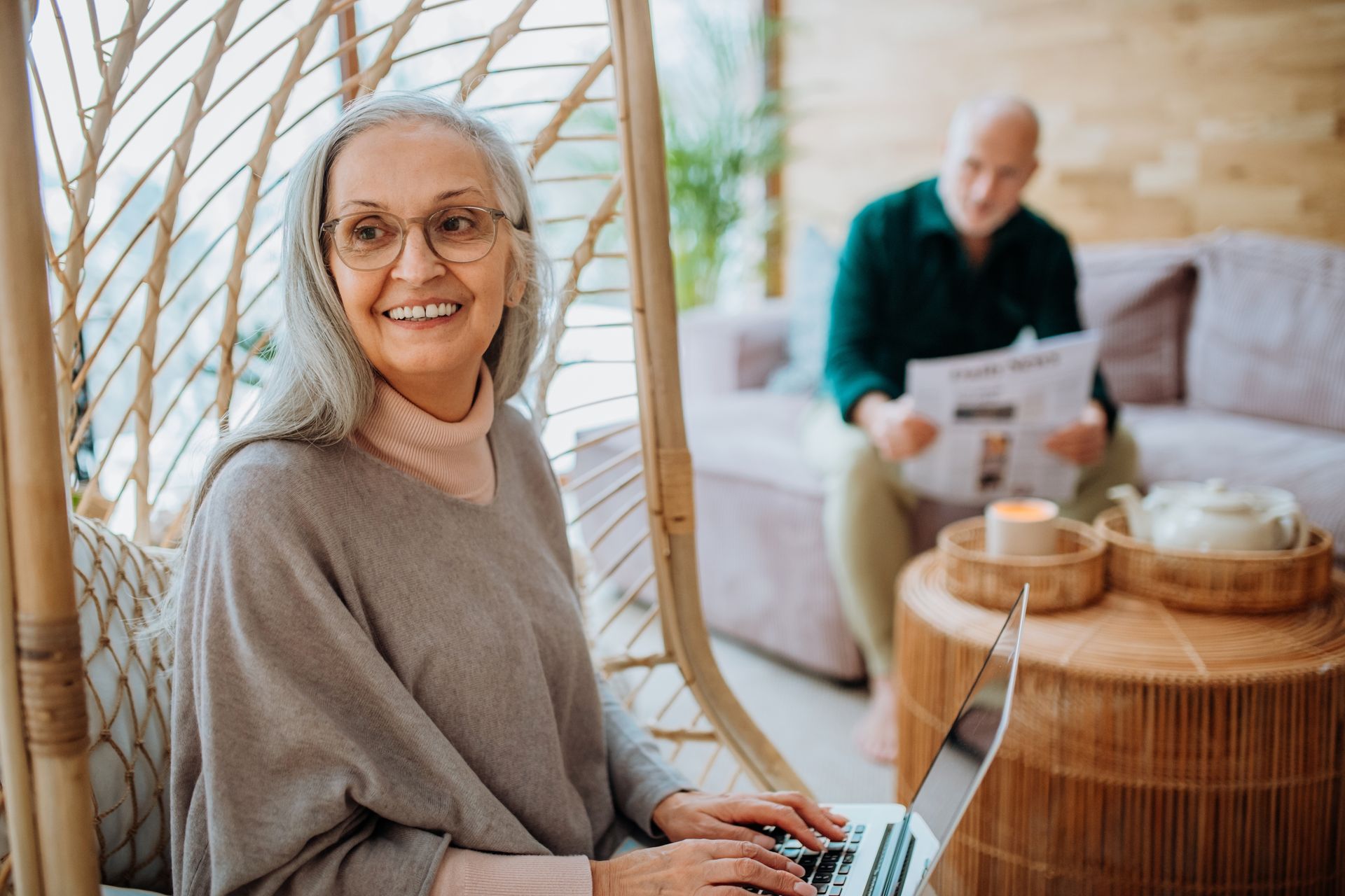 Woman with gray hair uses laptop, smiling. Man reads newspaper on couch in background.