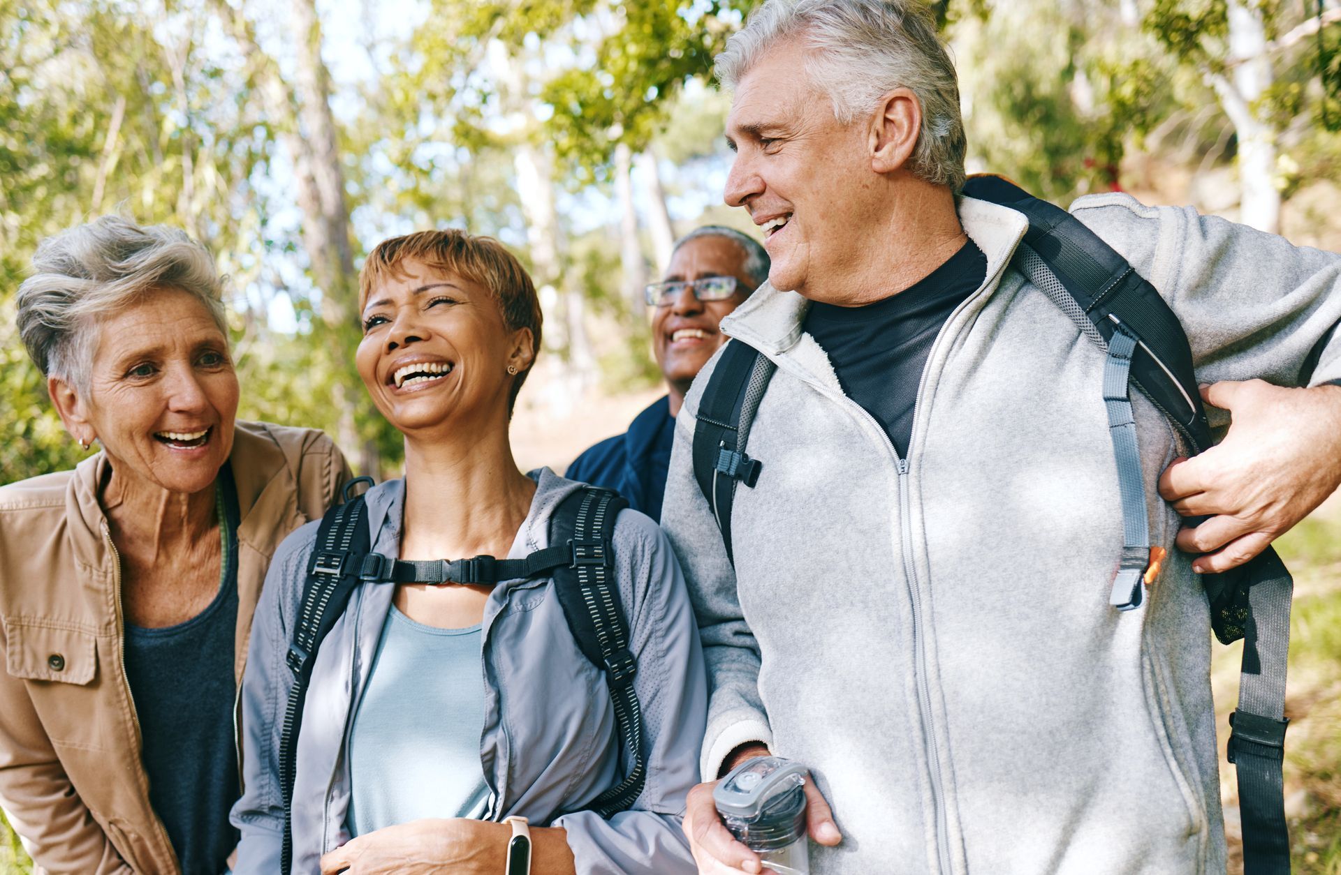 Four smiling older adults hiking in a sunny wooded area. They wear backpacks, and two are laughing.