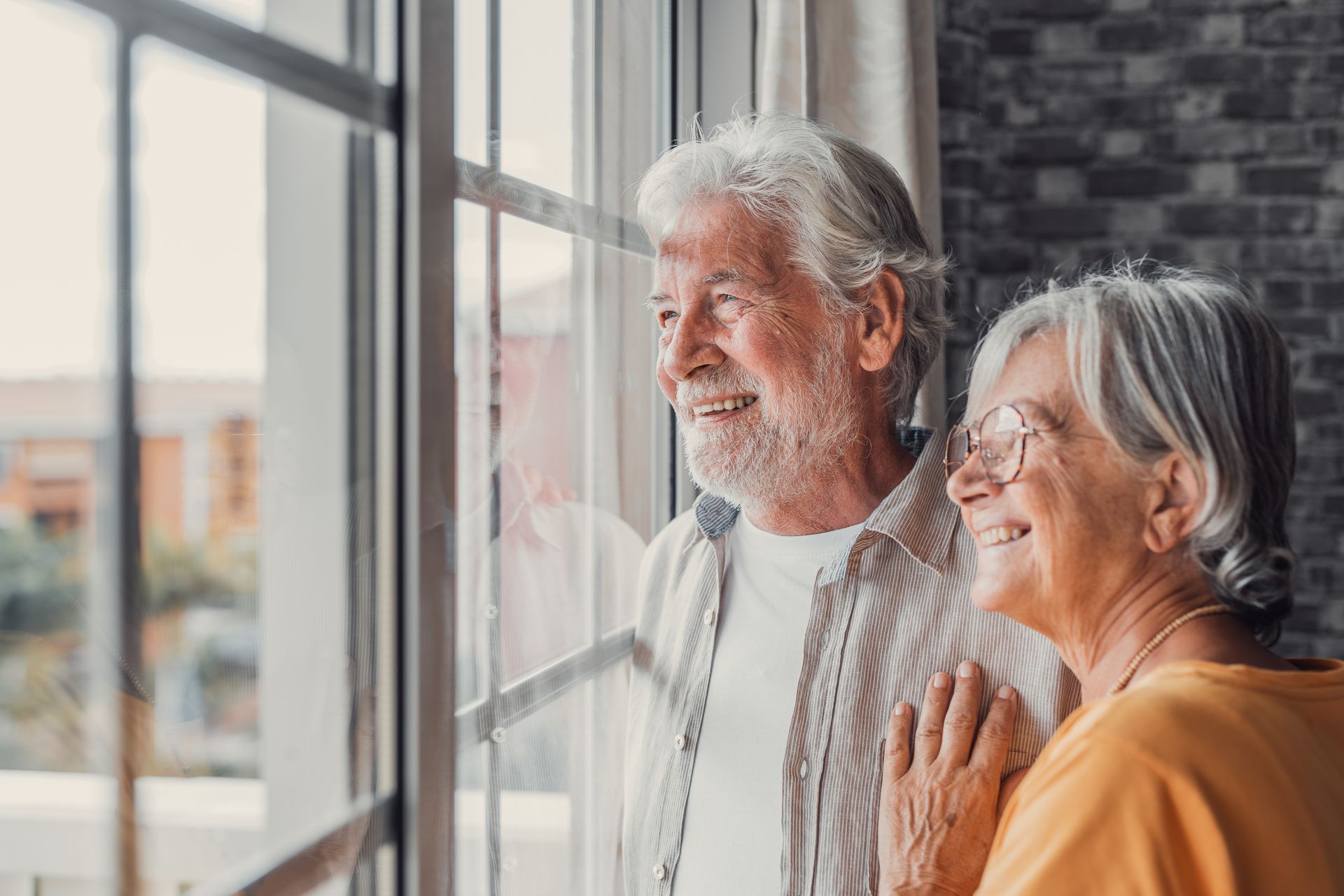 Elderly couple smiling and looking out a window, enjoying the view, inside a home.