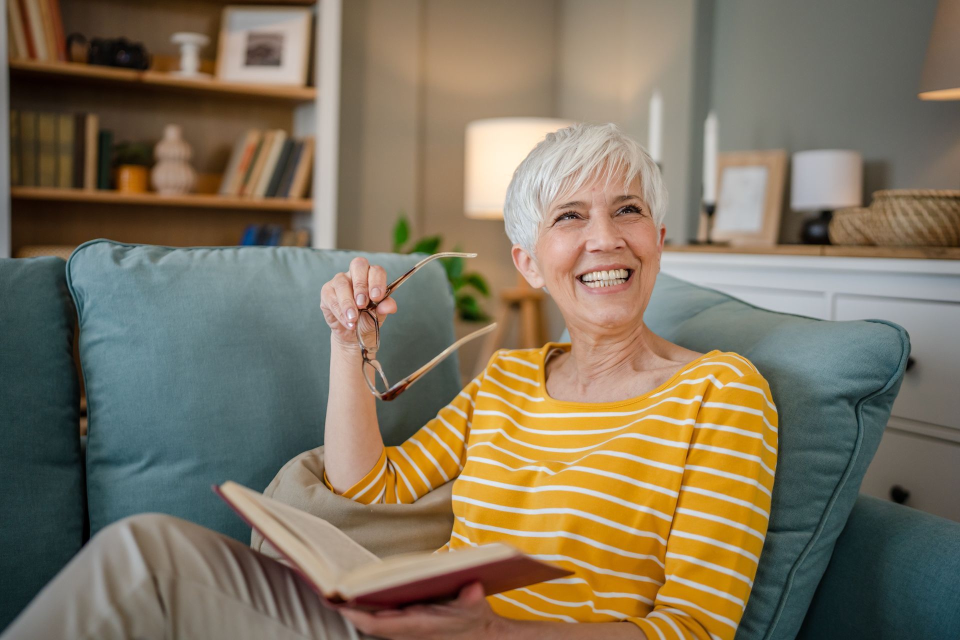 Smiling older woman in yellow striped shirt holding book and glasses, relaxing on a blue couch in a living room.
