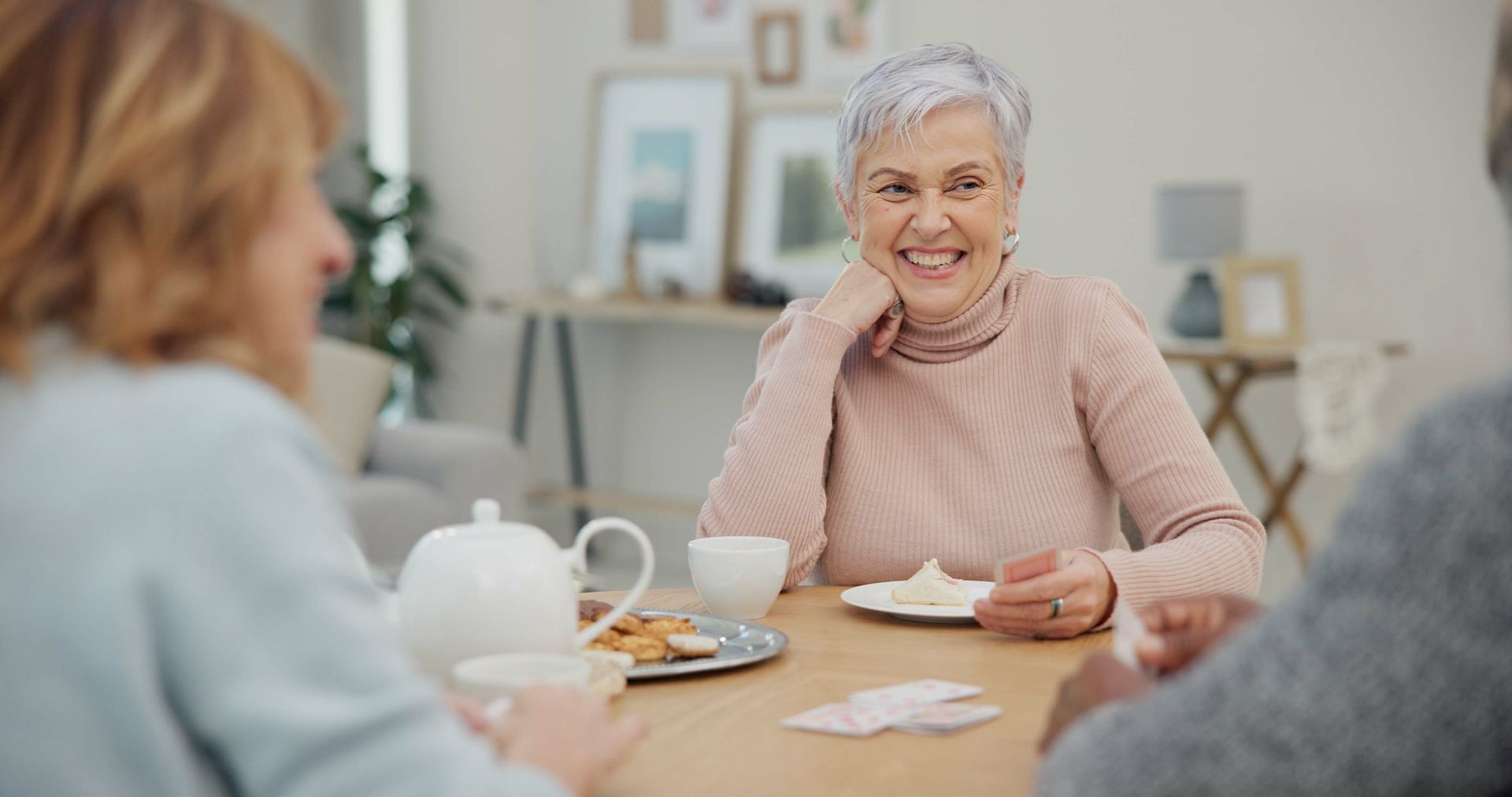 Three senior women enjoy a social gathering at a table. One smiles holding food, tea and cookies.
