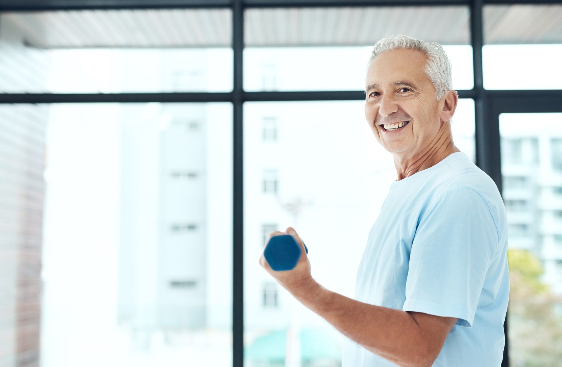 Smiling older man in a light blue shirt lifting a small blue dumbbell in front of a window.