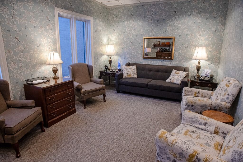 A furnished waiting room with floral wallpaper, sofa, armchairs, lamps, and a window.