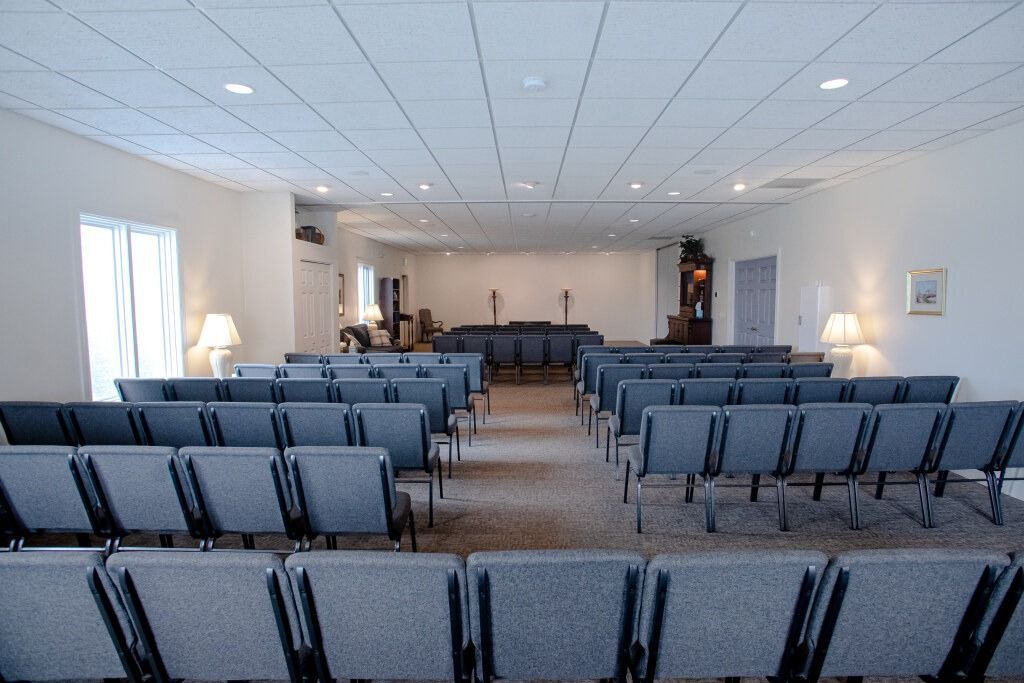Rows of blue chairs face a raised area, inside a light-filled room.