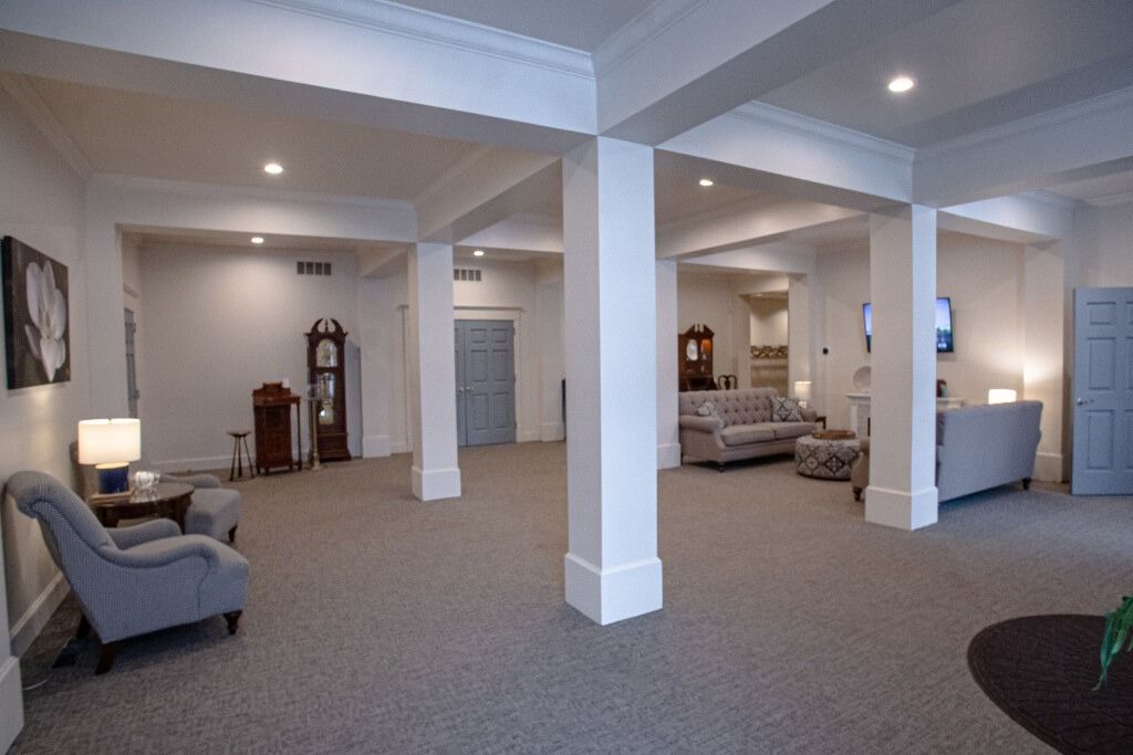 Spacious room with columns, carpet, sofas, chairs, and a grandfather clock. White walls, soft lighting.