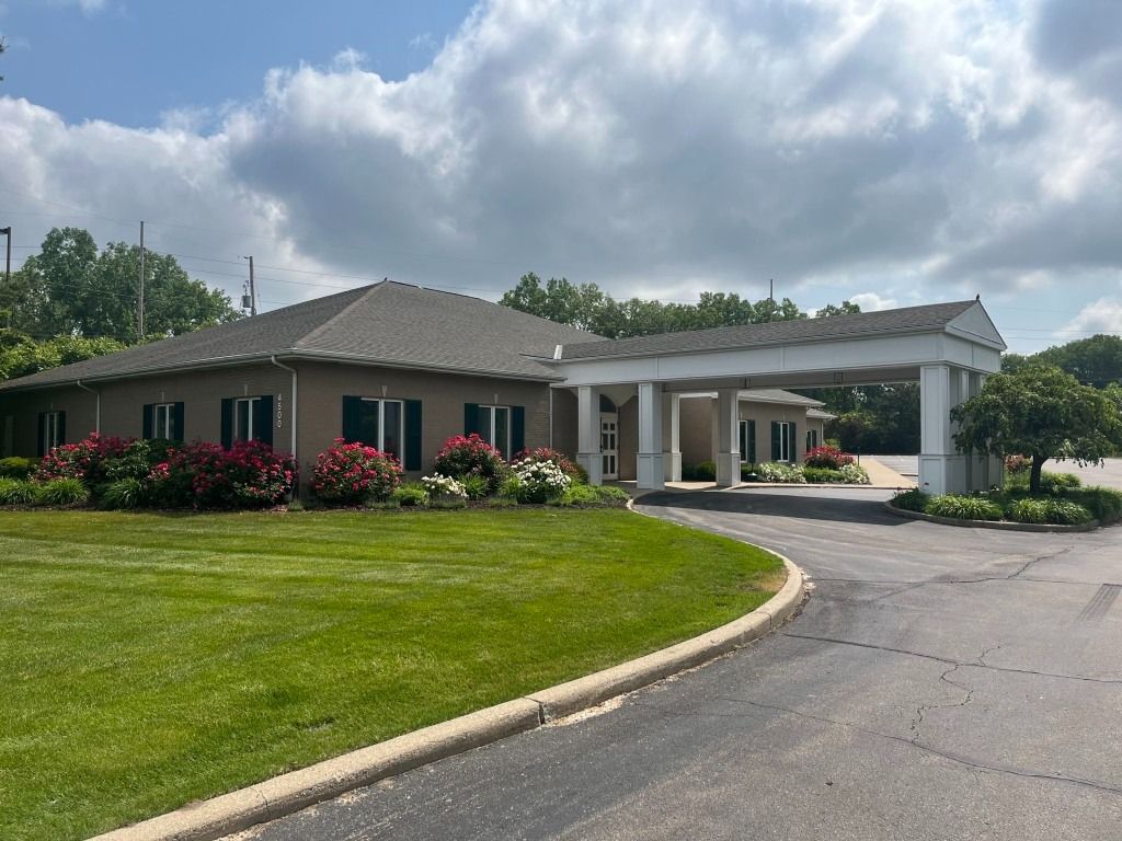 A one-story building with a covered entrance, green grass, flowers, and a cloudy sky.