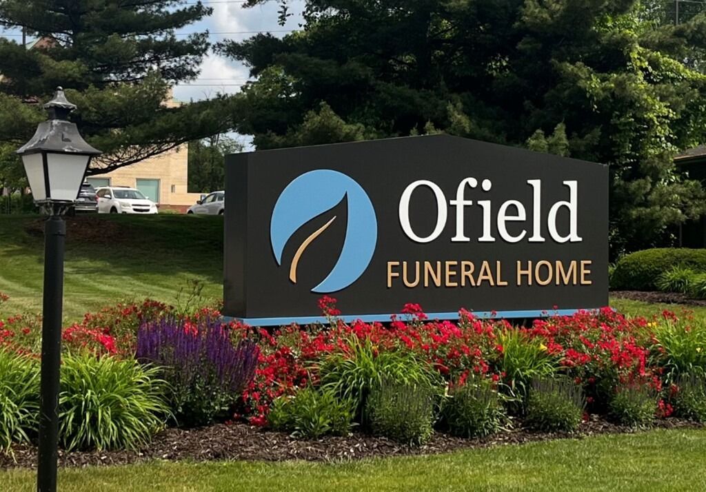 Ofield Funeral Home sign with blue and green leaf logo, surrounded by flowers, in front of a building.