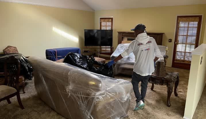A man is standing in a living room with a couch wrapped in plastic.