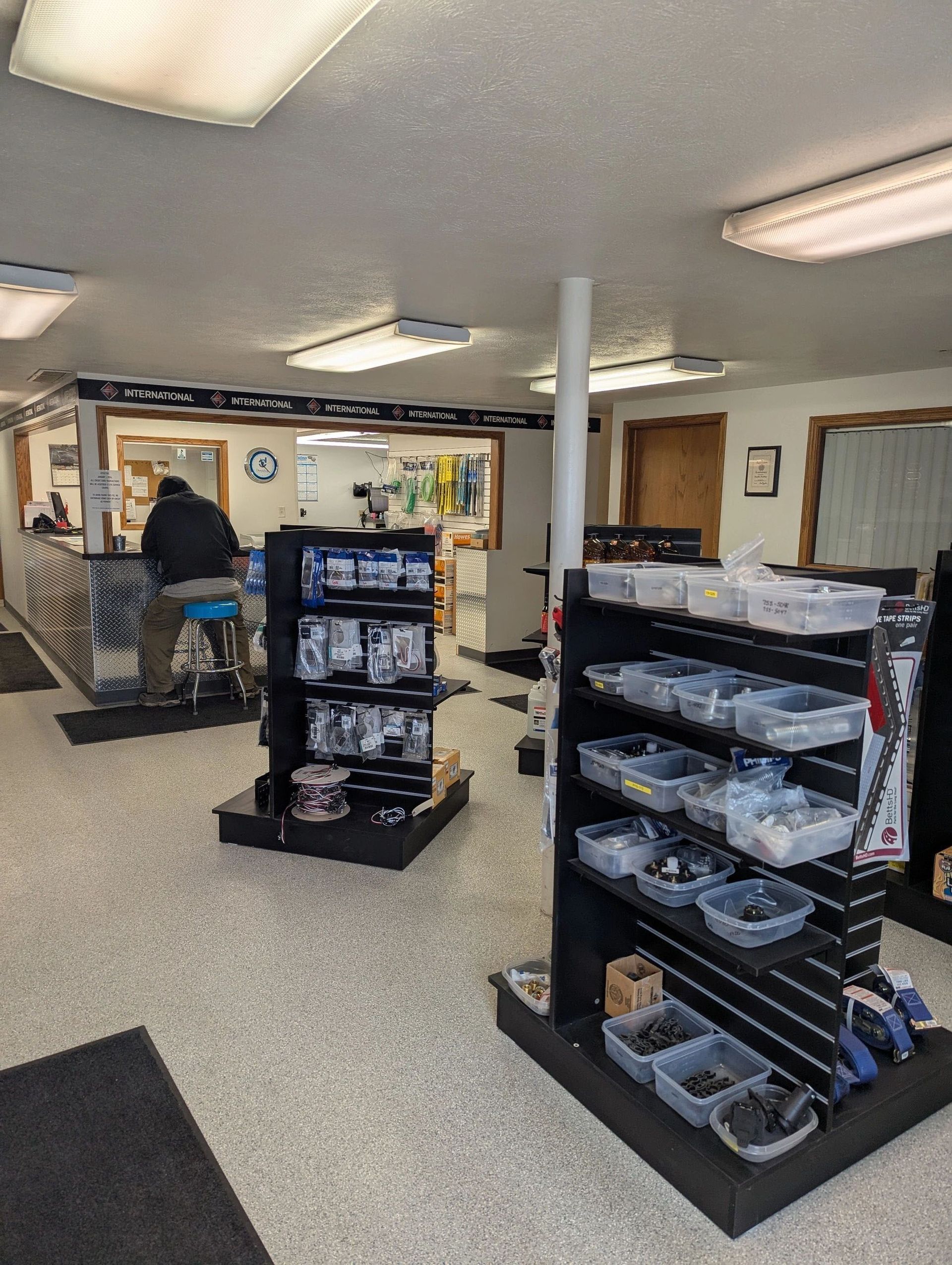 Inside of a hardware store, with shelves of items, customer at counter, and neutral-colored flooring.