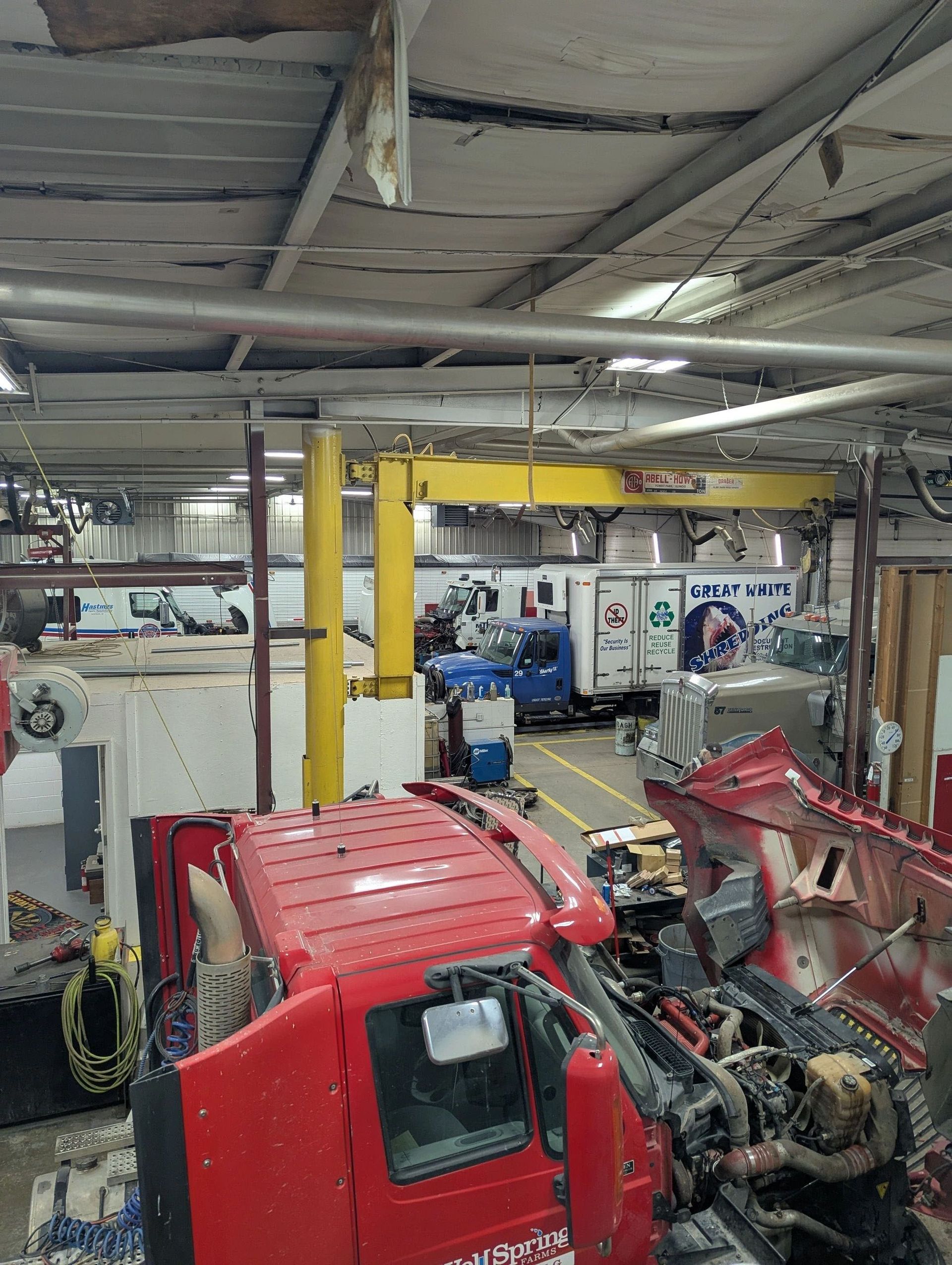 Red truck being worked on in a large garage.  A yellow crane and several other trucks are visible in the background.