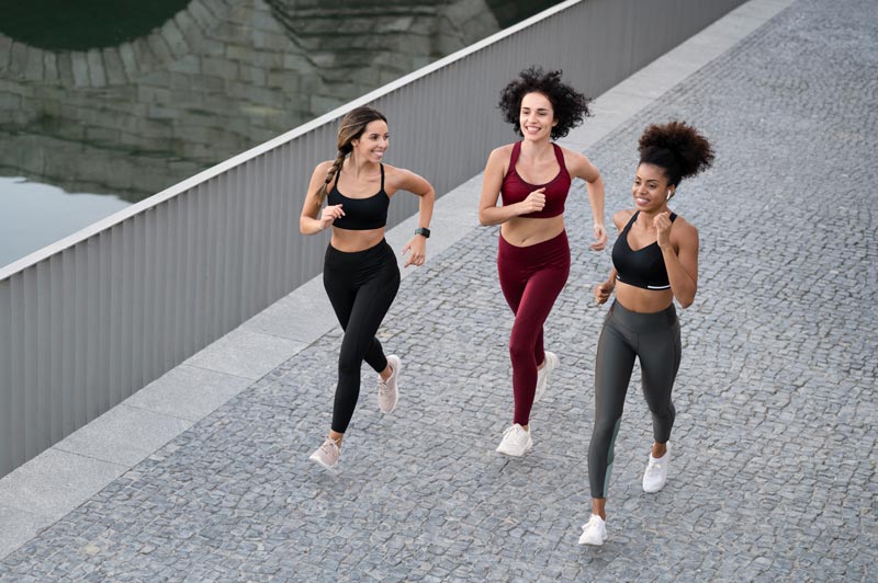 Three women are running on a sidewalk next to a body of water.