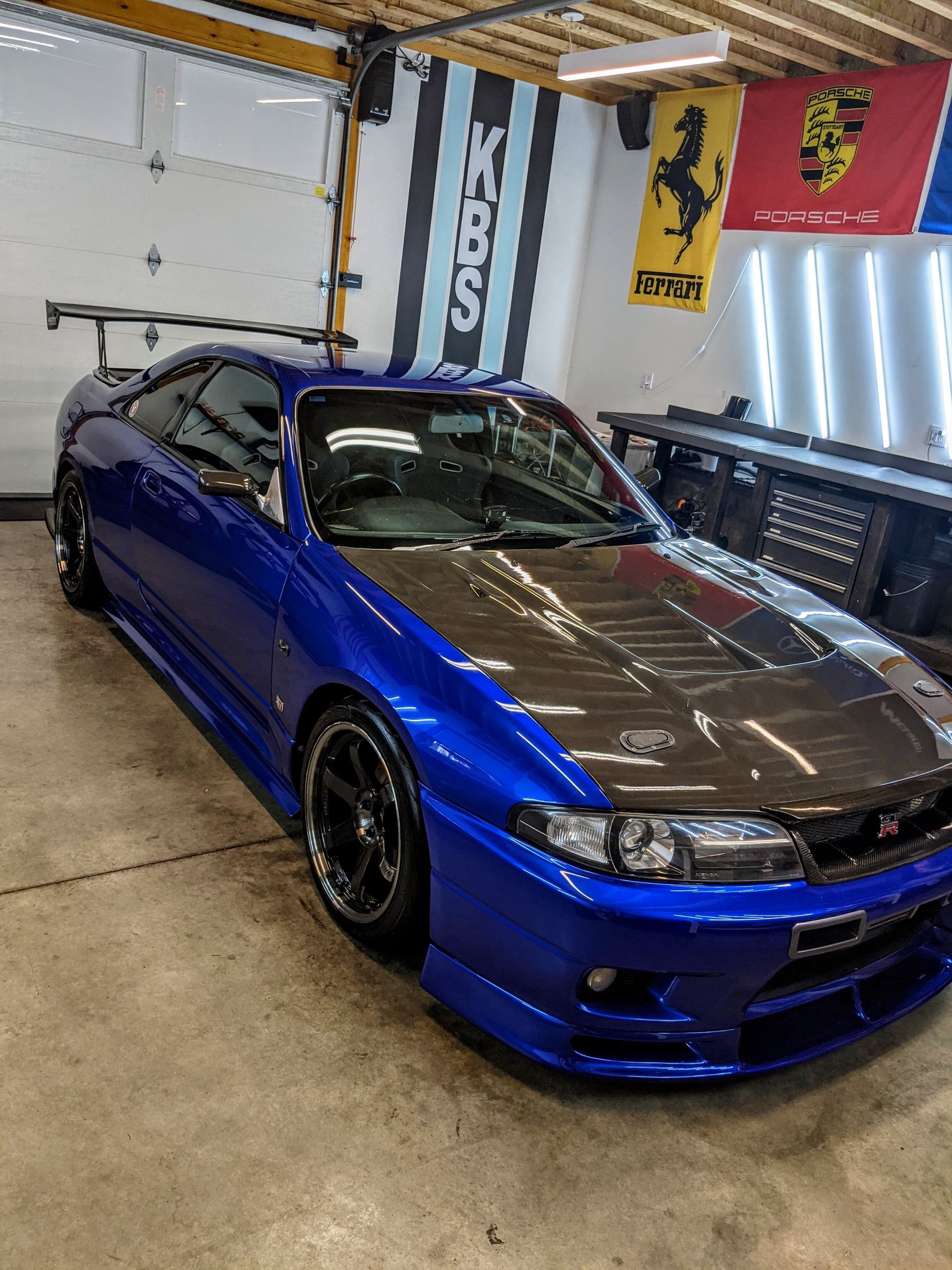 Blue sports coupe with carbon-fiber hood parked in a garage next to racing flags and lights