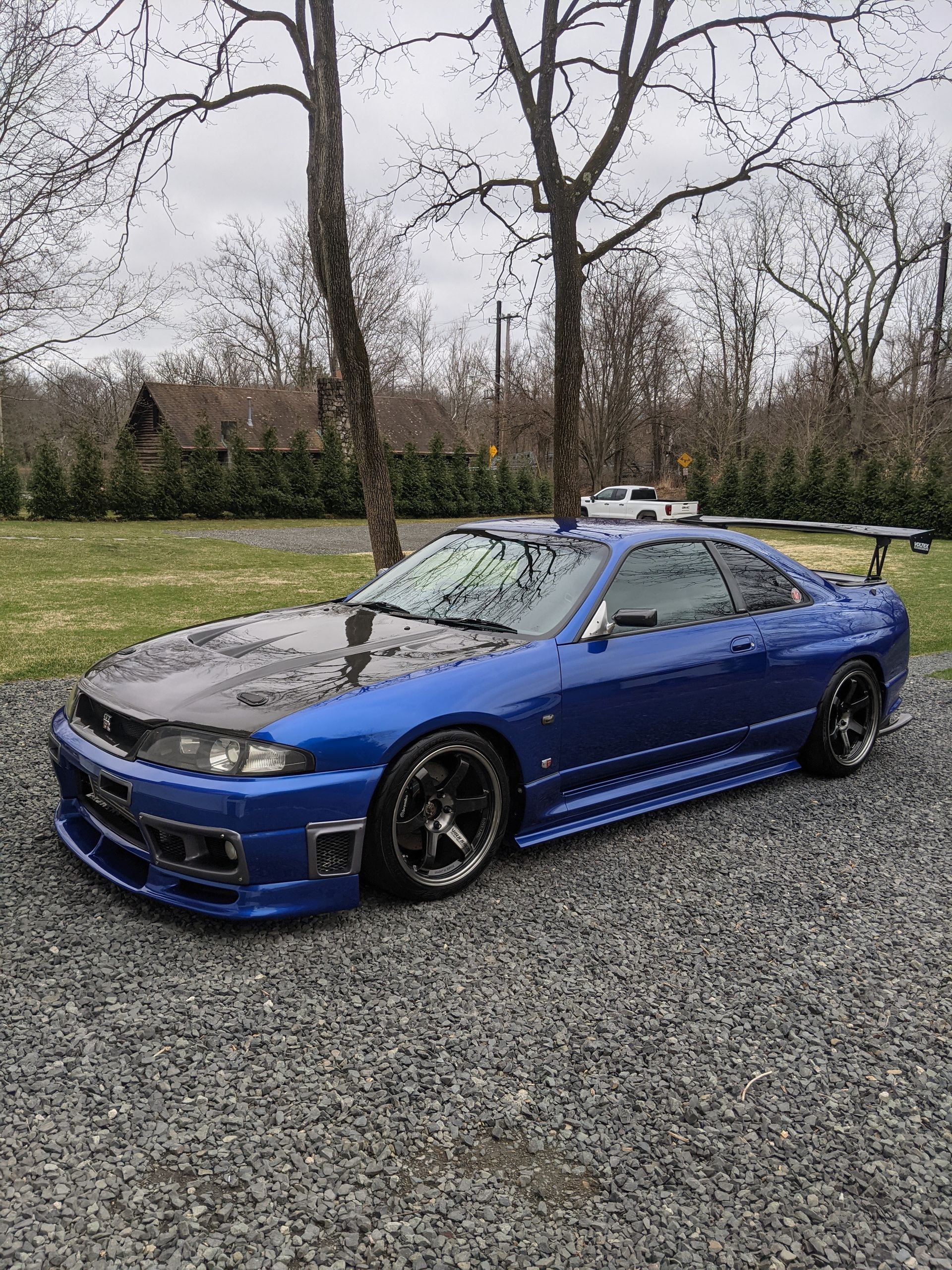 Blue sports coupe parked on gravel with a black hood and spoiler, outdoors by bare trees.