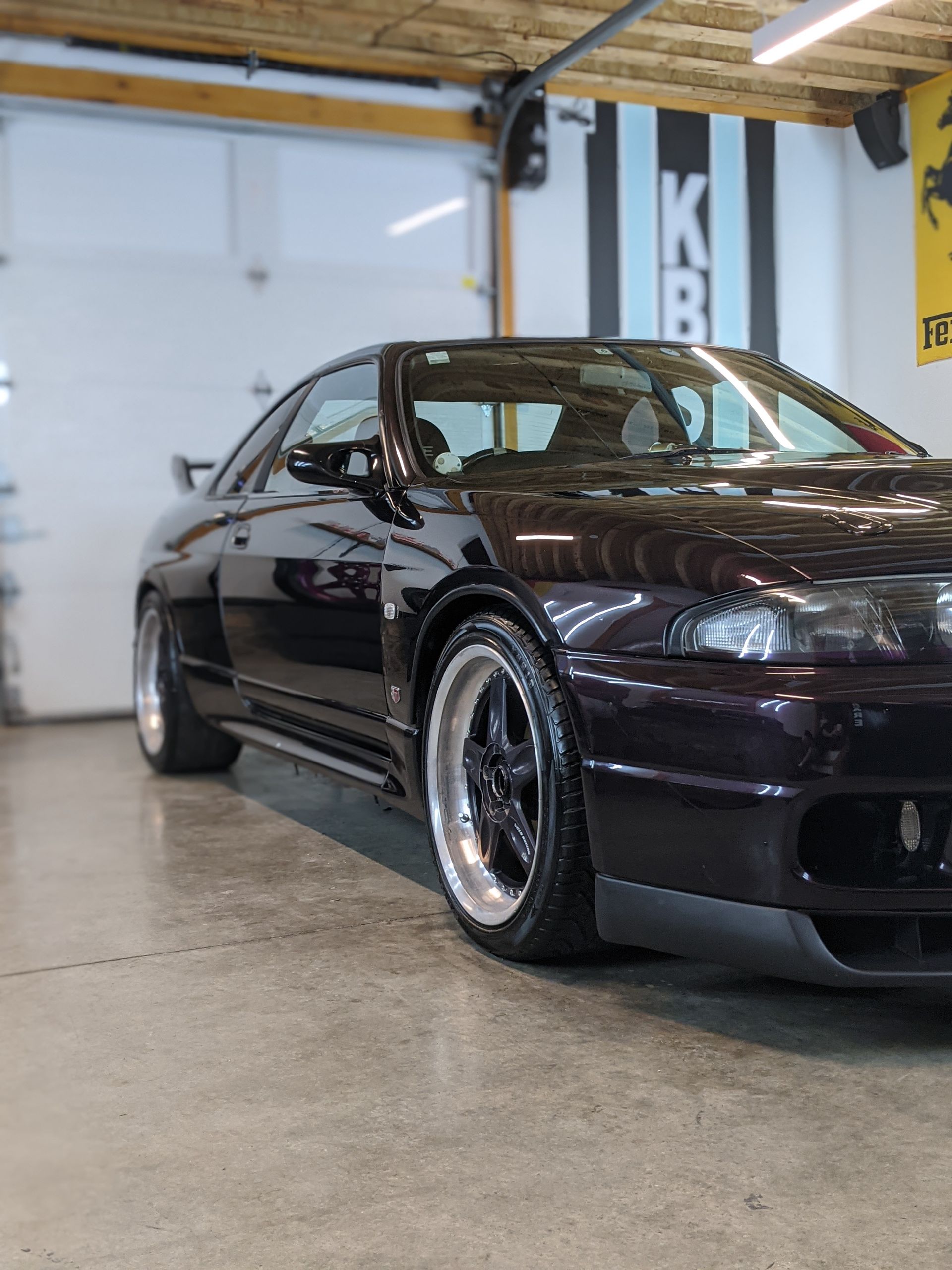 Black modified sports coupe in a garage, shown from the front-left side with polished wheels.