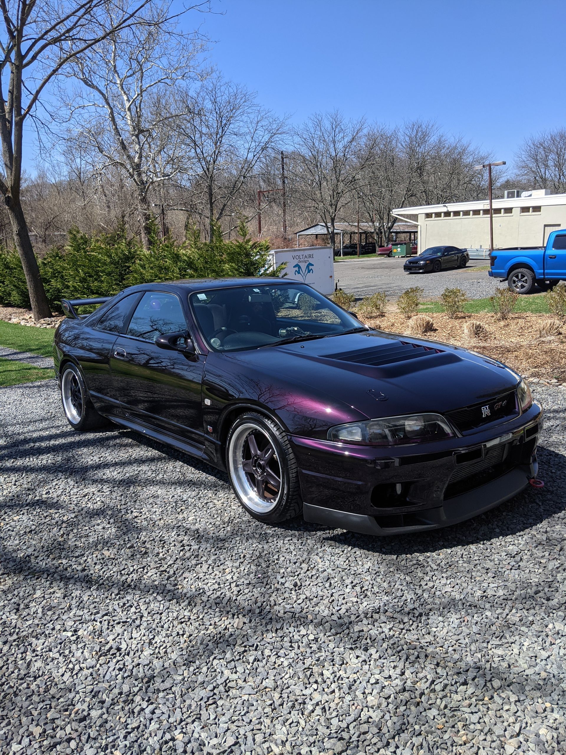 Black coupe parked on gravel with purple front fender, outdoors near trees and buildings.