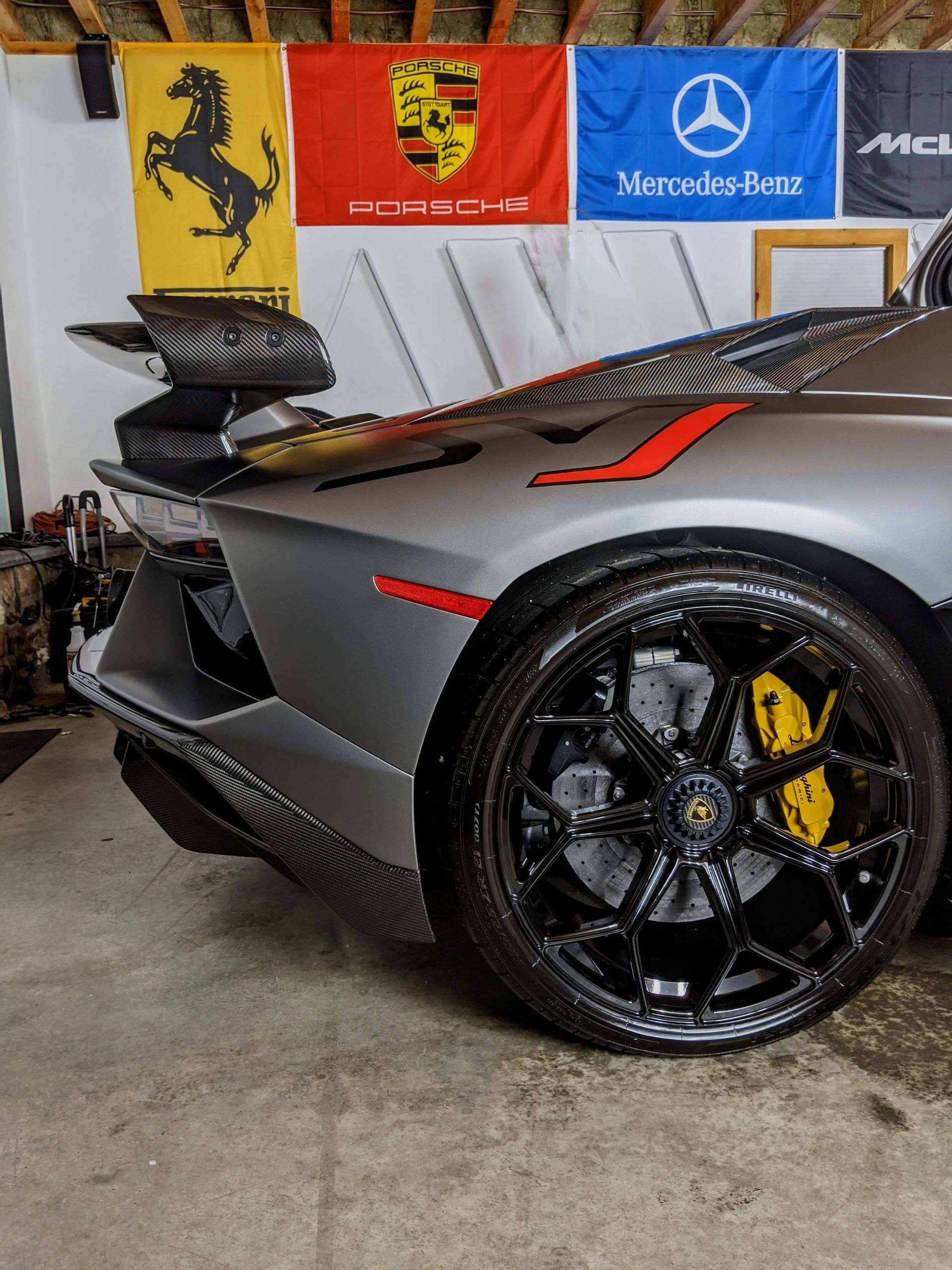 Gray sports car with black rims and yellow brake calipers in a garage with Ferrari and Mercedes flags