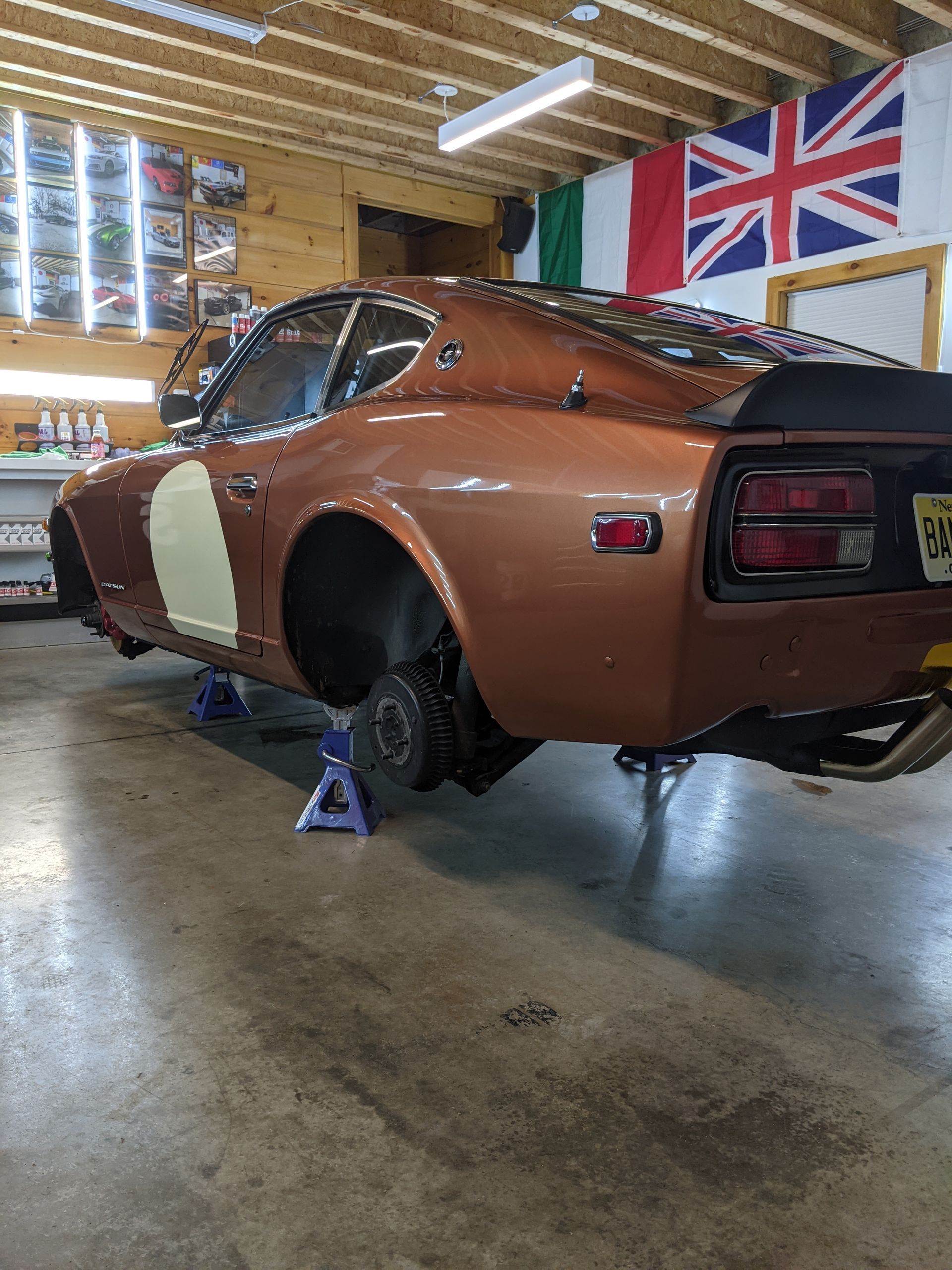 Brown classic muscle car on jack stands in a garage, with rear wheel removed and UK flags hanging nearby