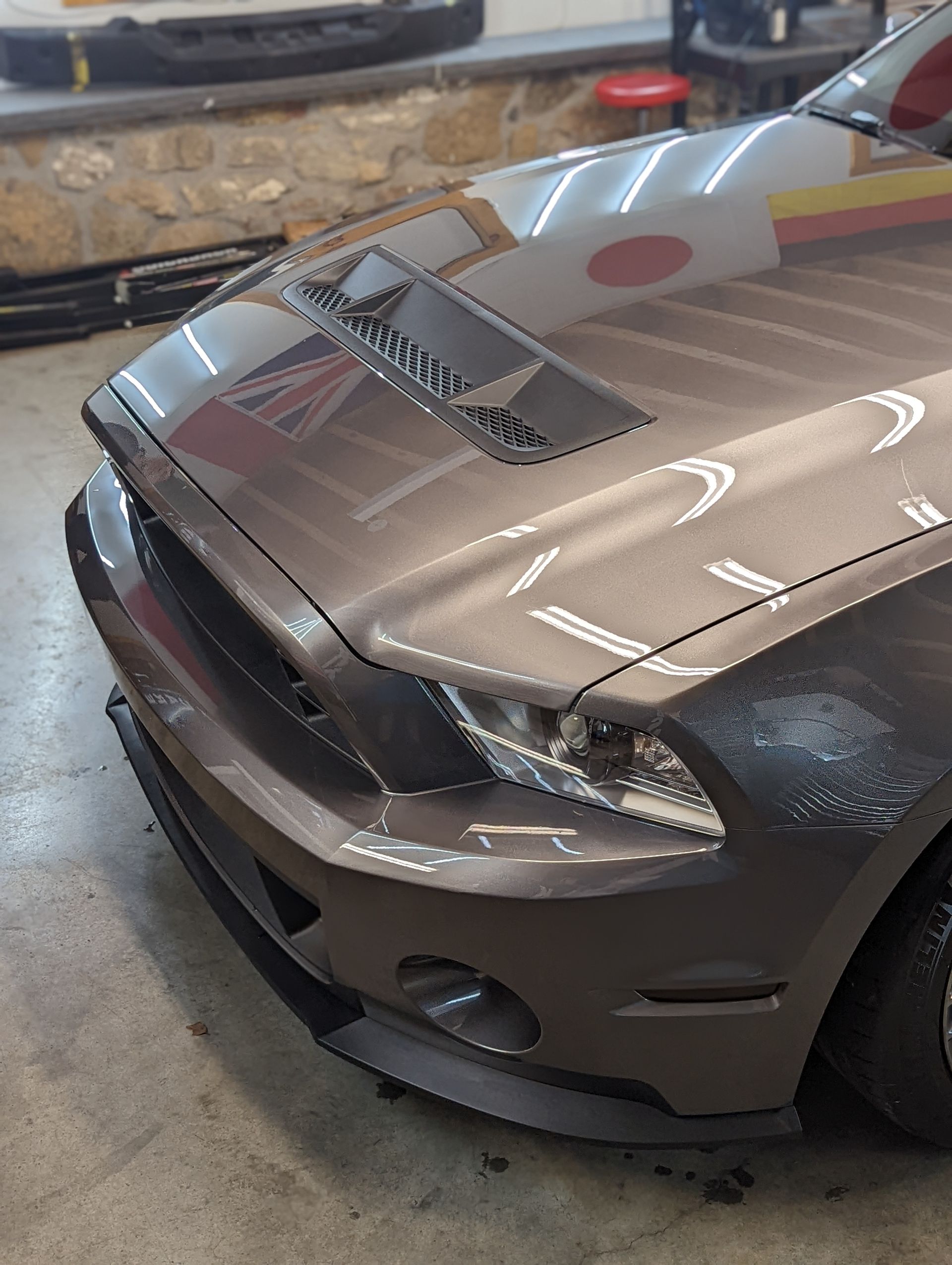Damaged tan car front-left hood and headlight in a garage, with visible dents and scratches