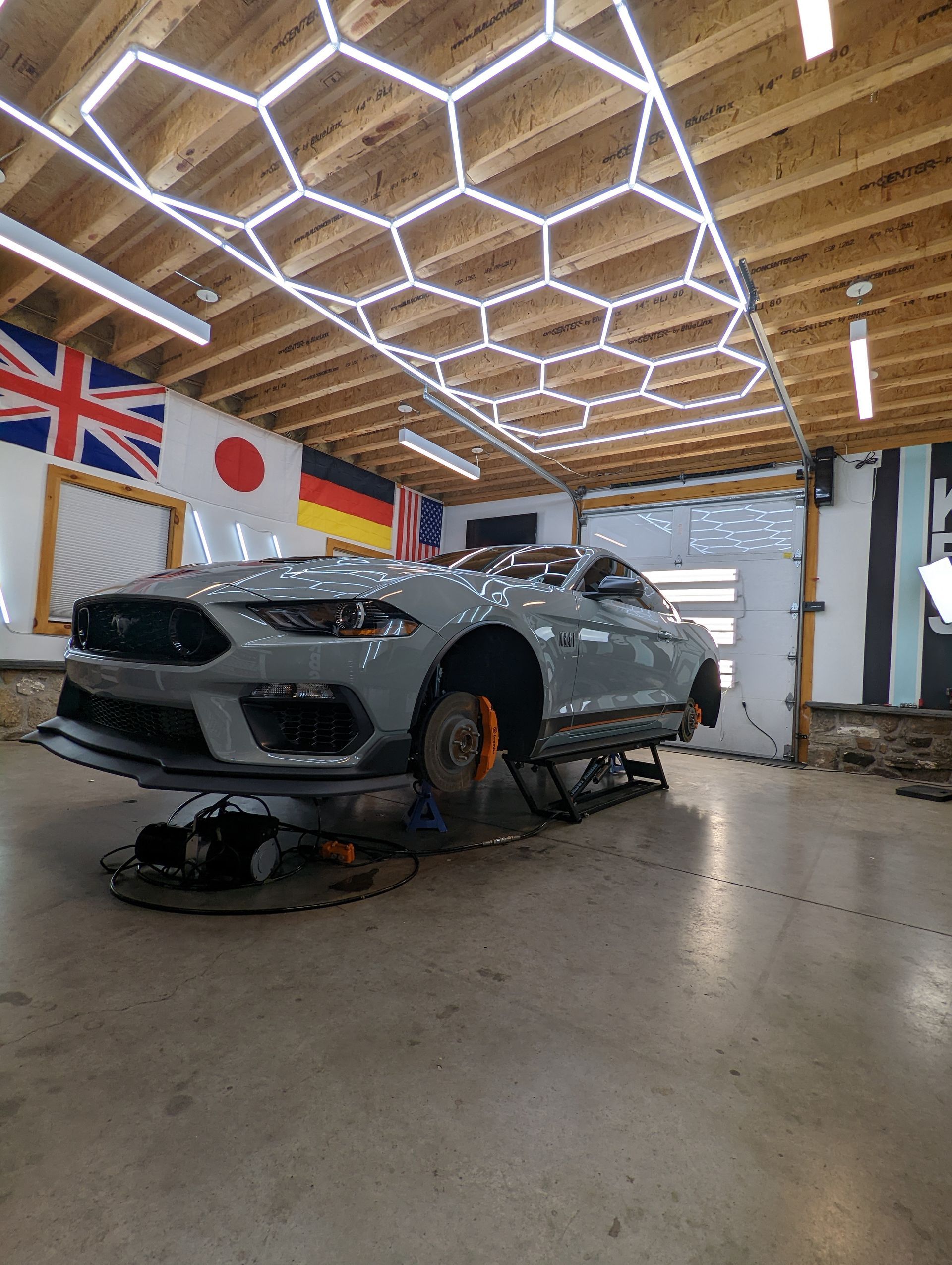 Grey sports car on a lift in a workshop with flags on the wall and hexagonal ceiling lights.
