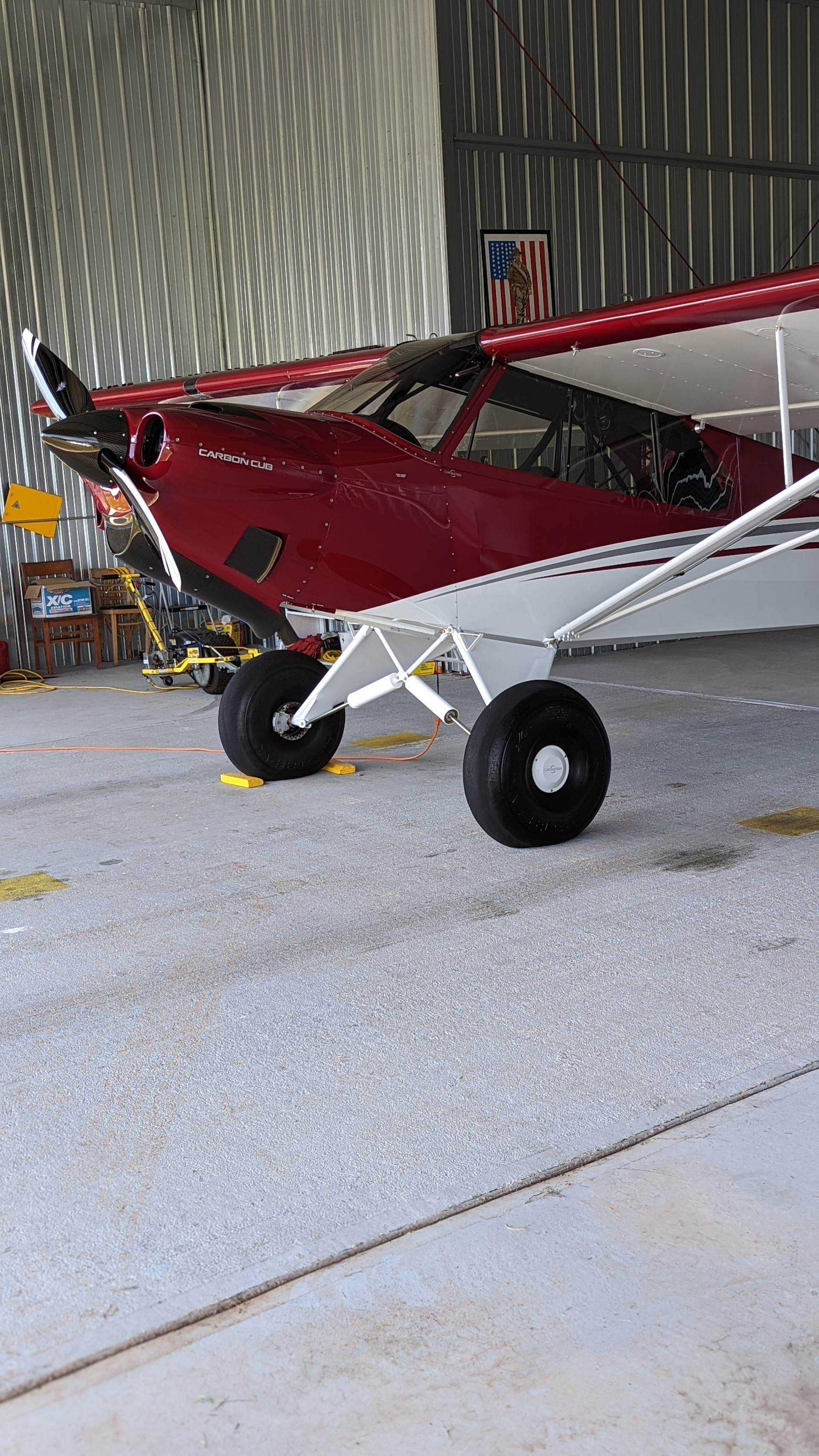 Red and white small airplane parked in a hangar with corrugated metal walls.