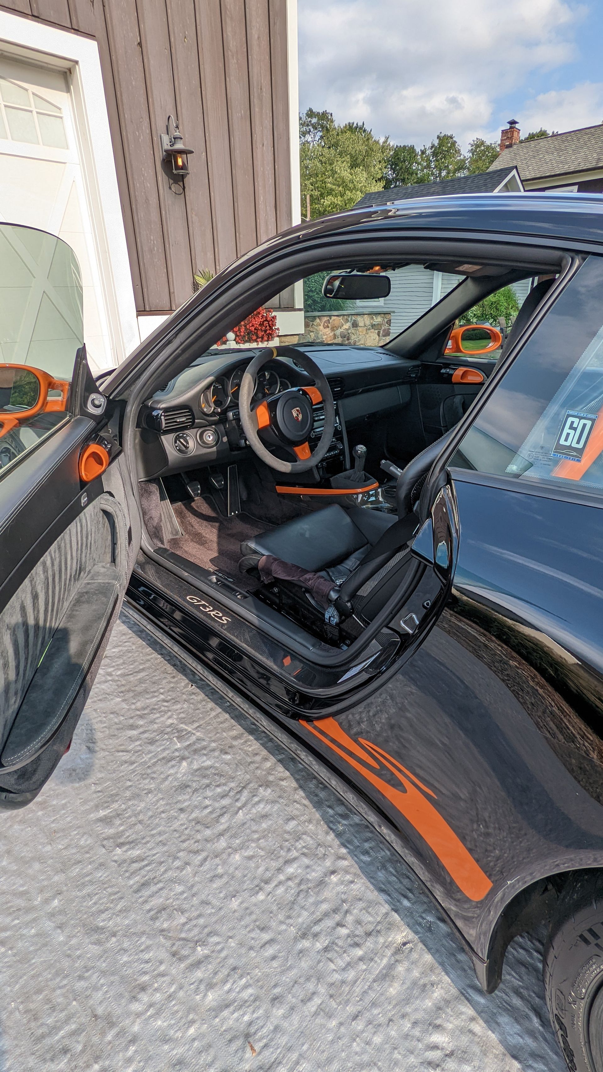 Black sports car door open, showing orange-accented steering wheel and interior beside a house.