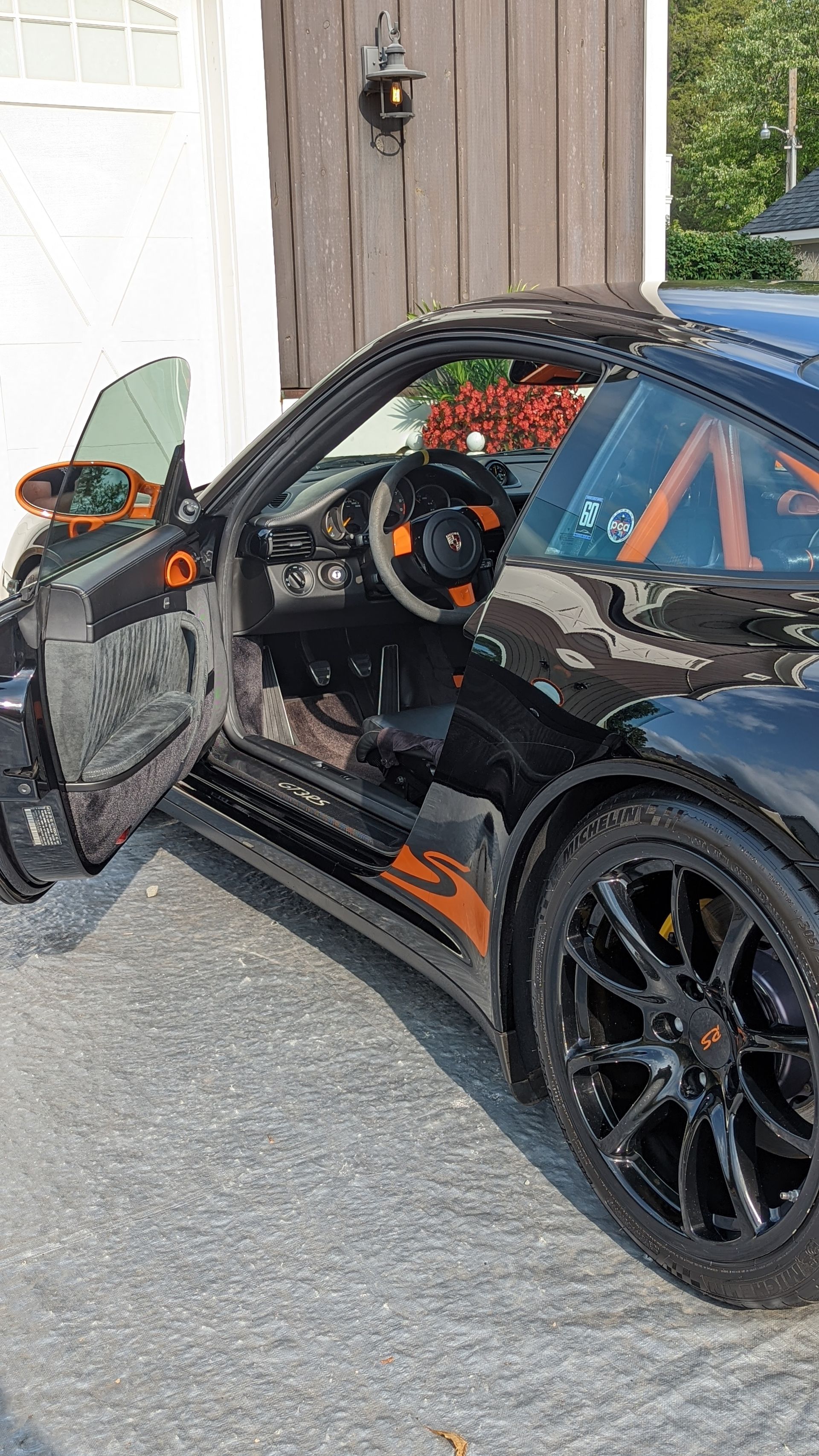 Black sports car with driver’s door open beside a garage, showing a black-and-orange interior.