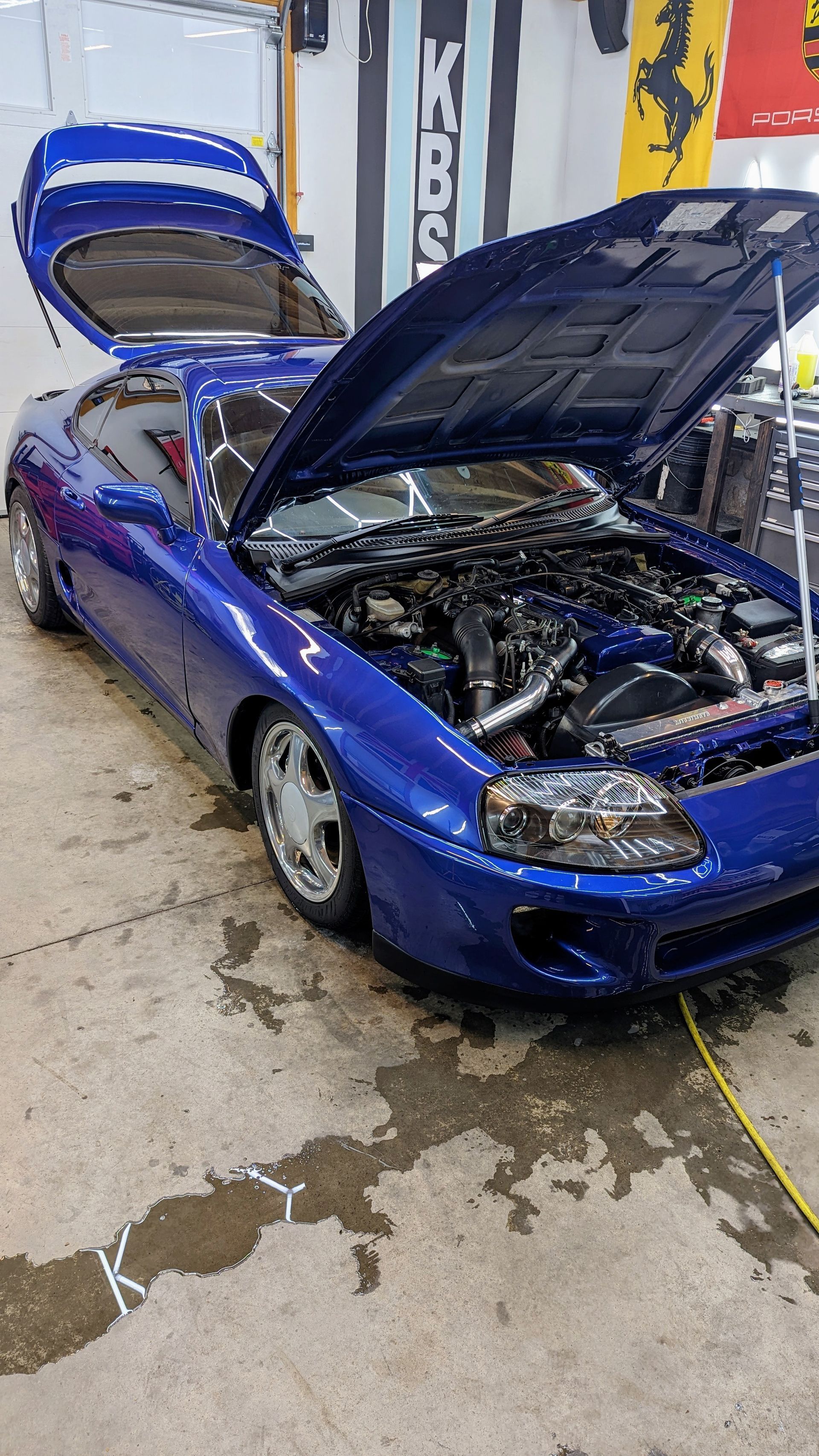 Blue sports car with hood and hatch open, displaying the engine in a garage.