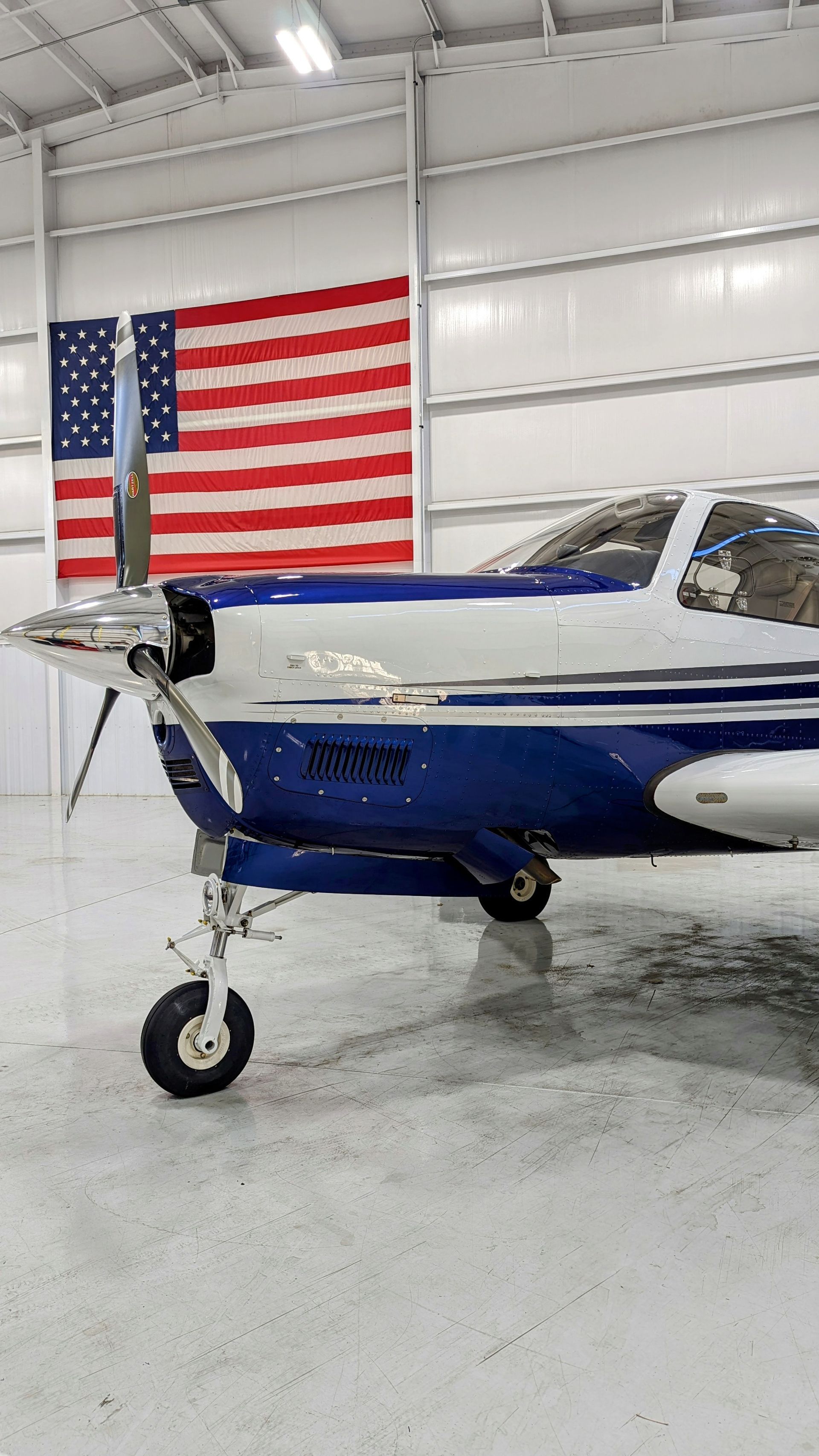 Blue-and-white small airplane inside a hangar with a large American flag on the wall