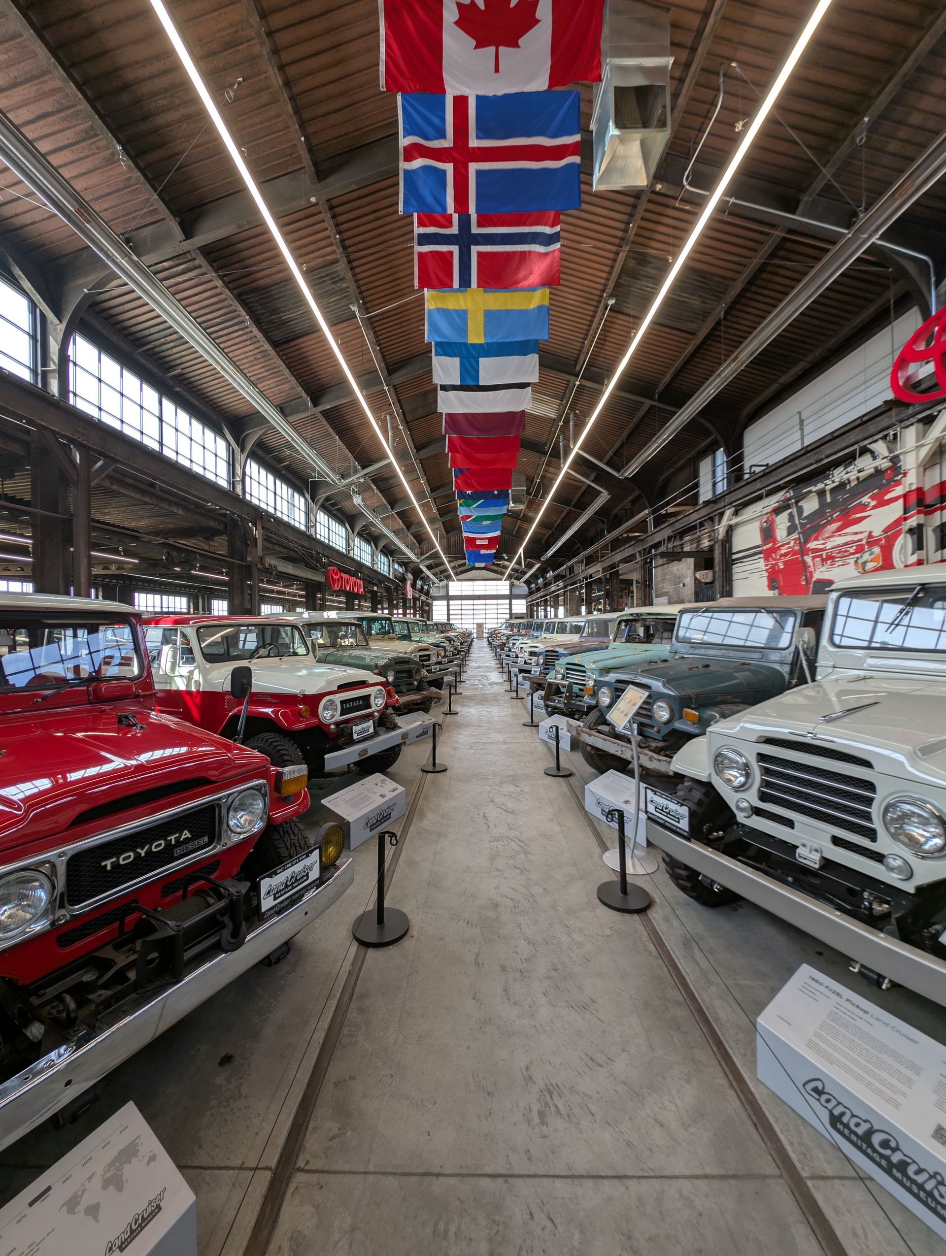 Classic cars lined up in a hall with flags hanging from the ceiling and a central aisle.