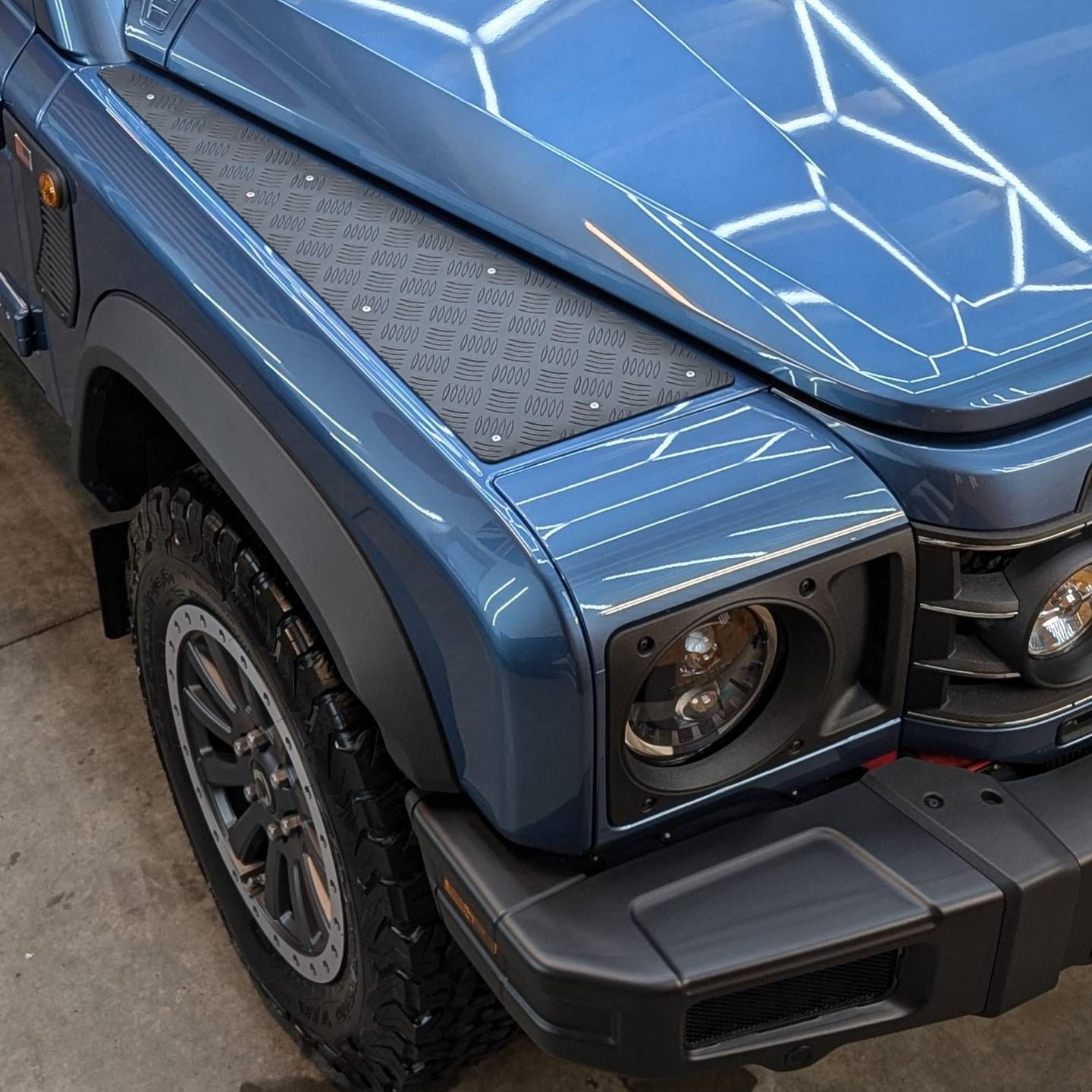 Close-up of a blue off-road SUV’s front left corner, showing headlight, tire, and bumper.