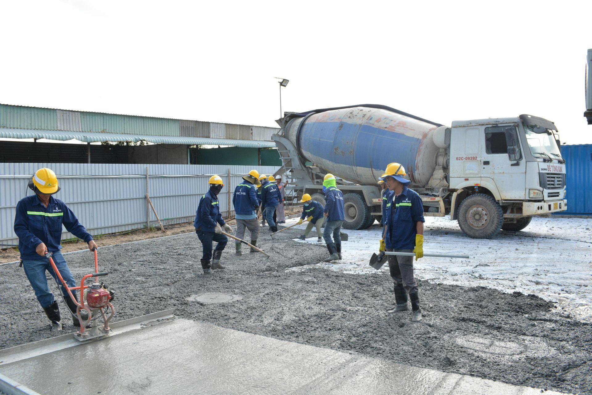 Construction workers paving a road beside a cement mixer truck, using yellow safety helmets