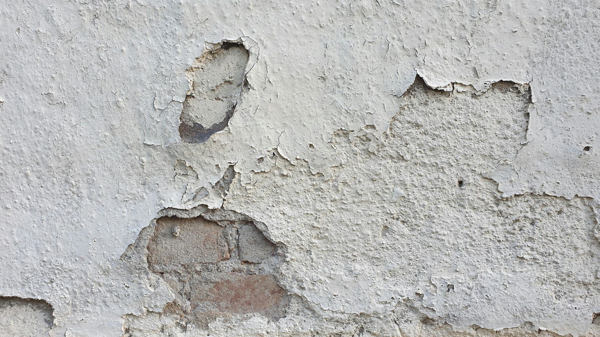 A weathered, gray stone wall with patches of plaster peeling away to reveal the rough brick and mortar beneath.