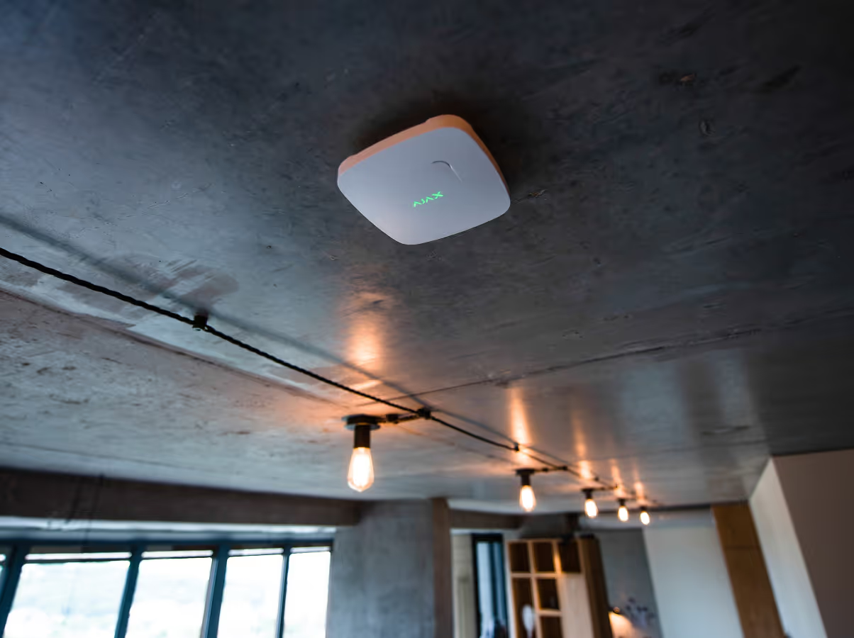 A White Fire Detector Mounted On A Dark, Industrial-style Concrete Ceiling Above A Row Of Hanging Pendant Lights — Cussler NQ in Airlie Beach, QLD