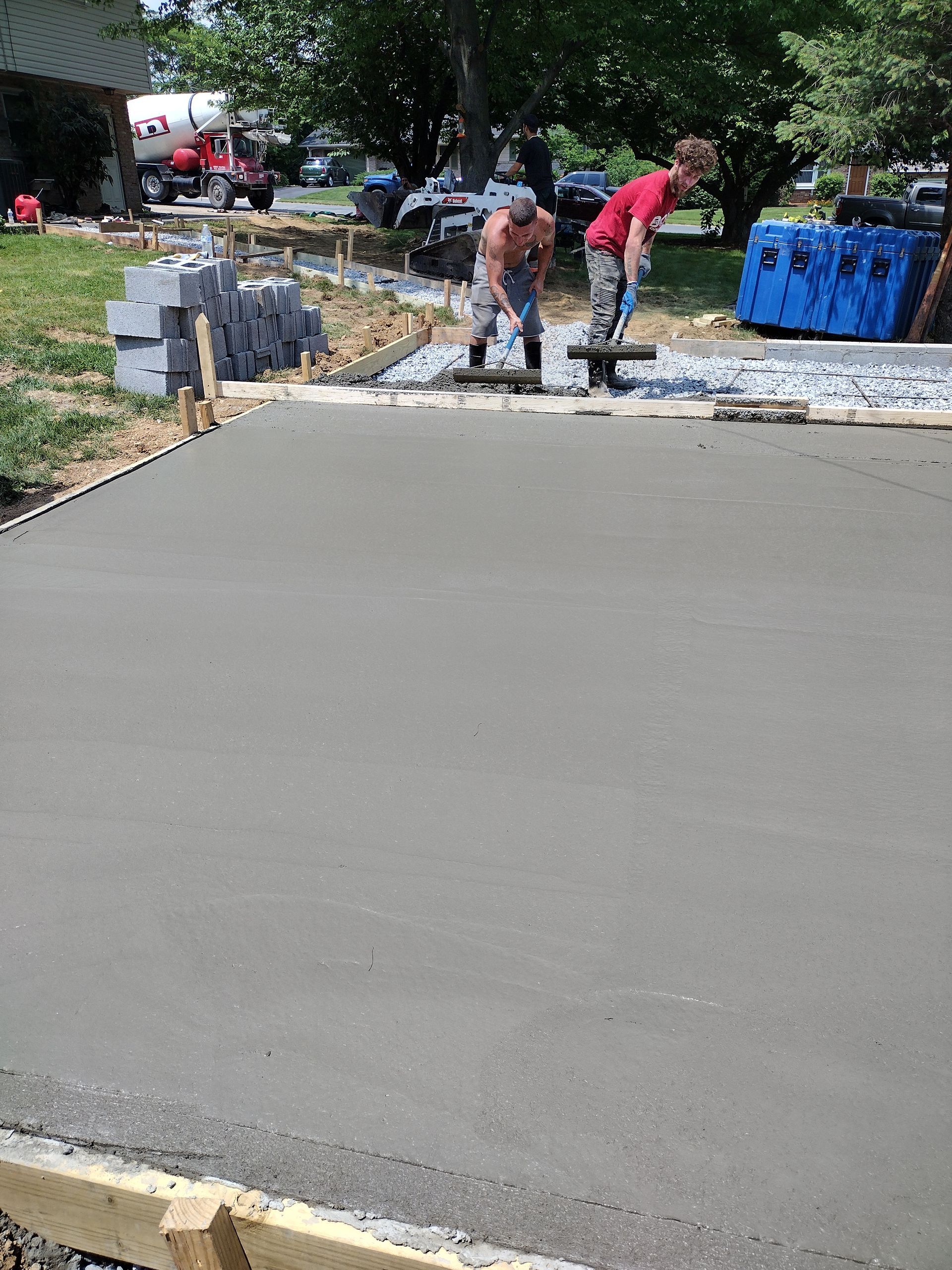 Two workers in safety gear finish leveling fresh gray concrete on a residential driveway construction site.