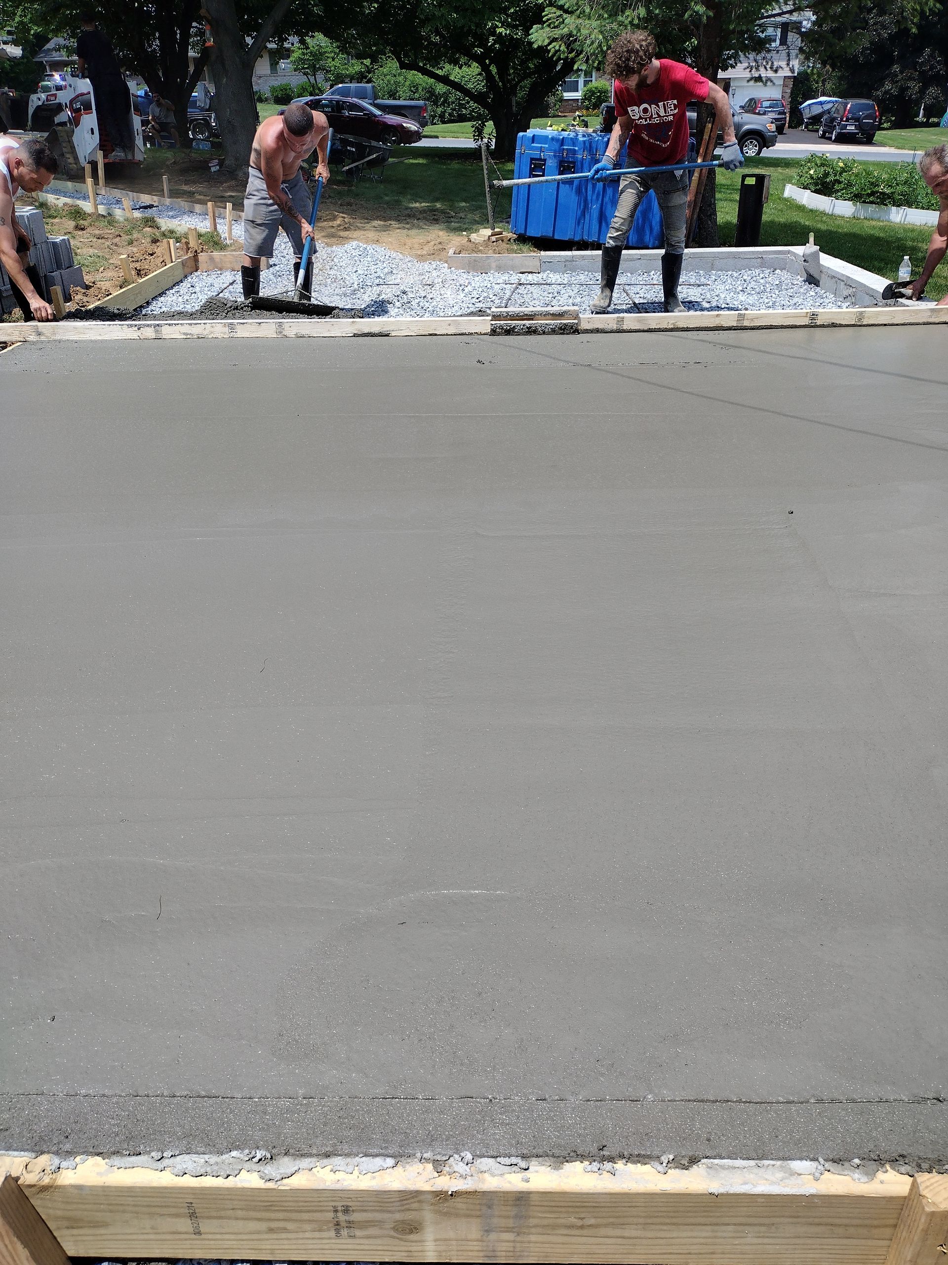 Two people in work clothes smoothing freshly poured concrete inside a wooden frame on a sunny day.