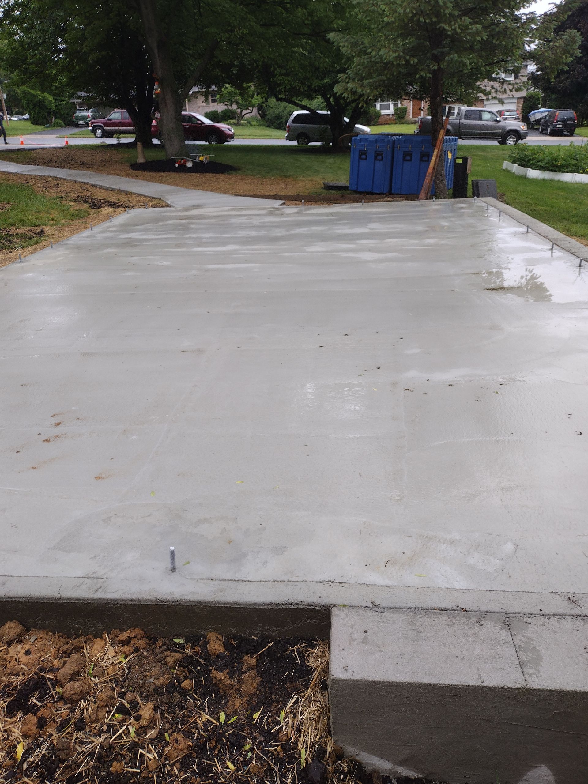 A newly poured rectangular concrete slab in a grassy yard, with a sidewalk leading to it and trees in the background.
