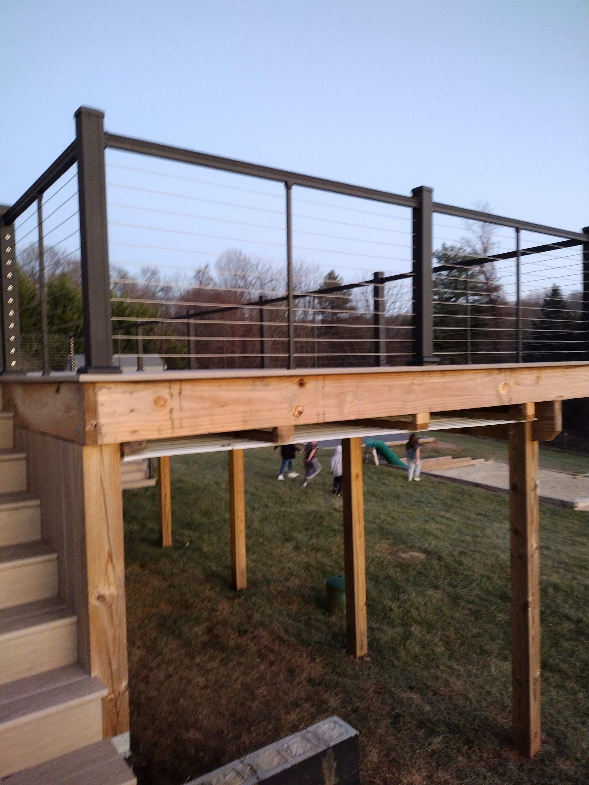 A wooden deck with a black metal cable railing stands above a grassy yard where people are playing.