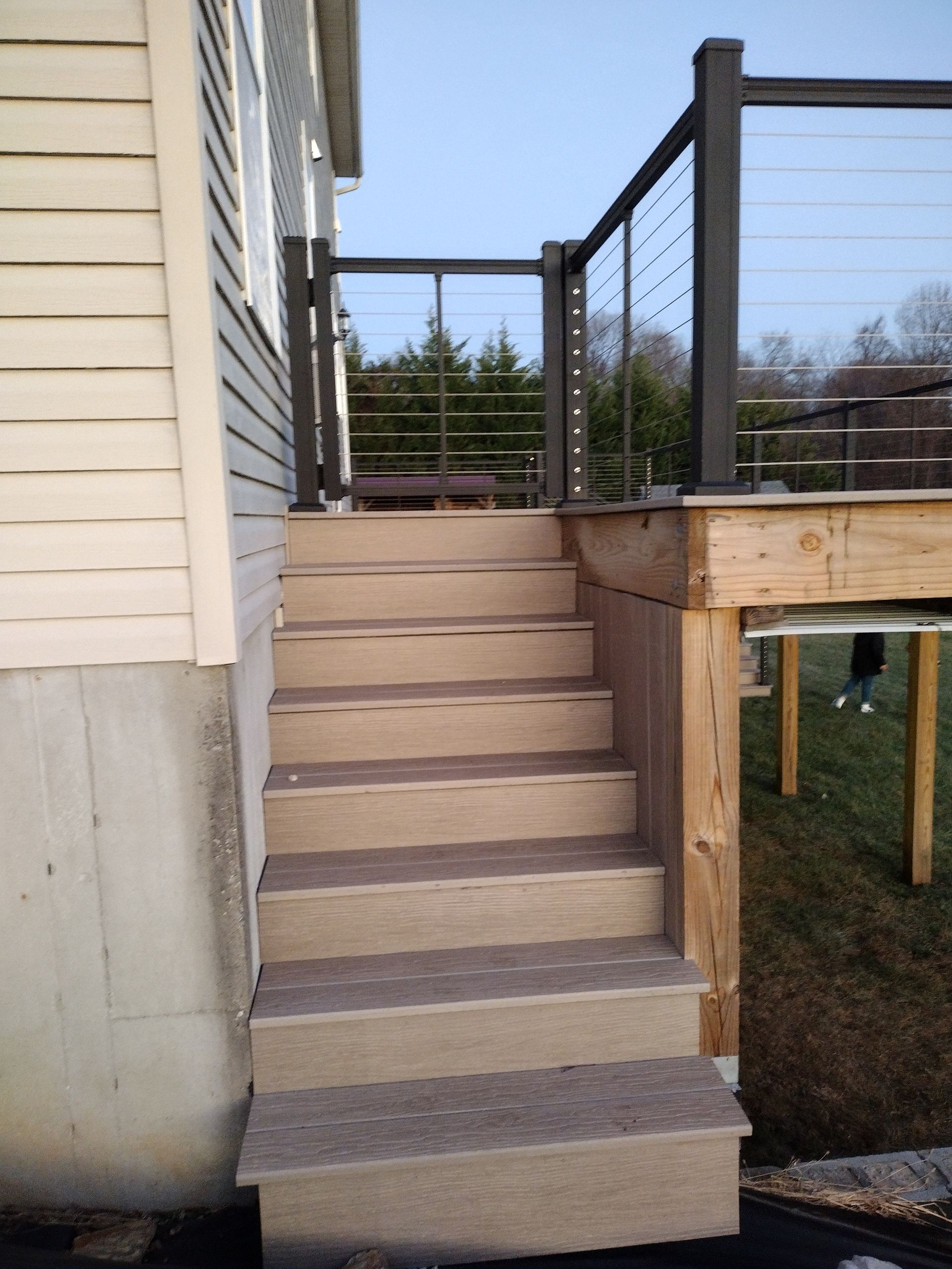A set of tan, synthetic stairs leading up to an elevated deck with dark railings against a beige house exterior.