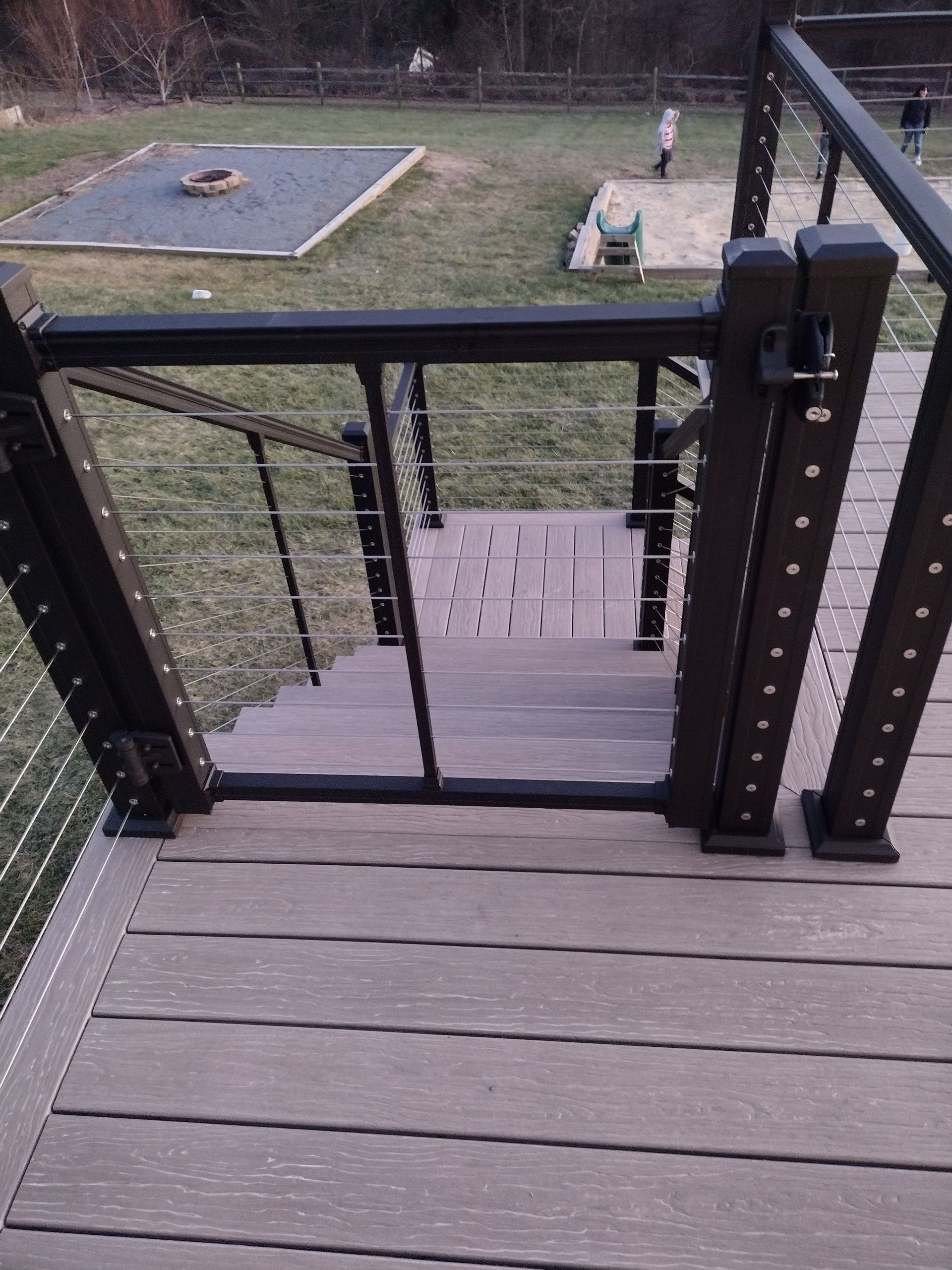 A black railing gate secures the top of a set of stairs on a wooden deck, overlooking a backyard lawn with a patio area.