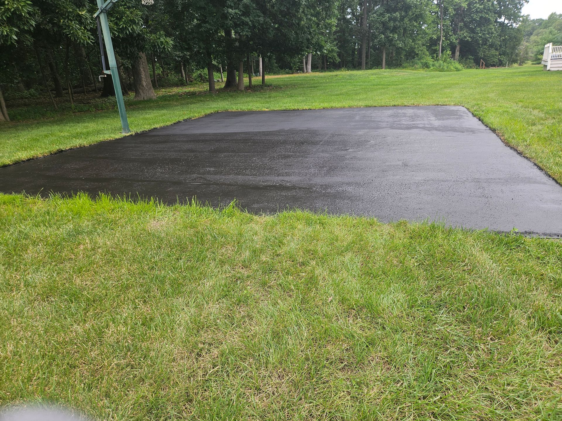 A newly paved black asphalt basketball court surface in a grassy backyard, with a basketball hoop pole on the left.